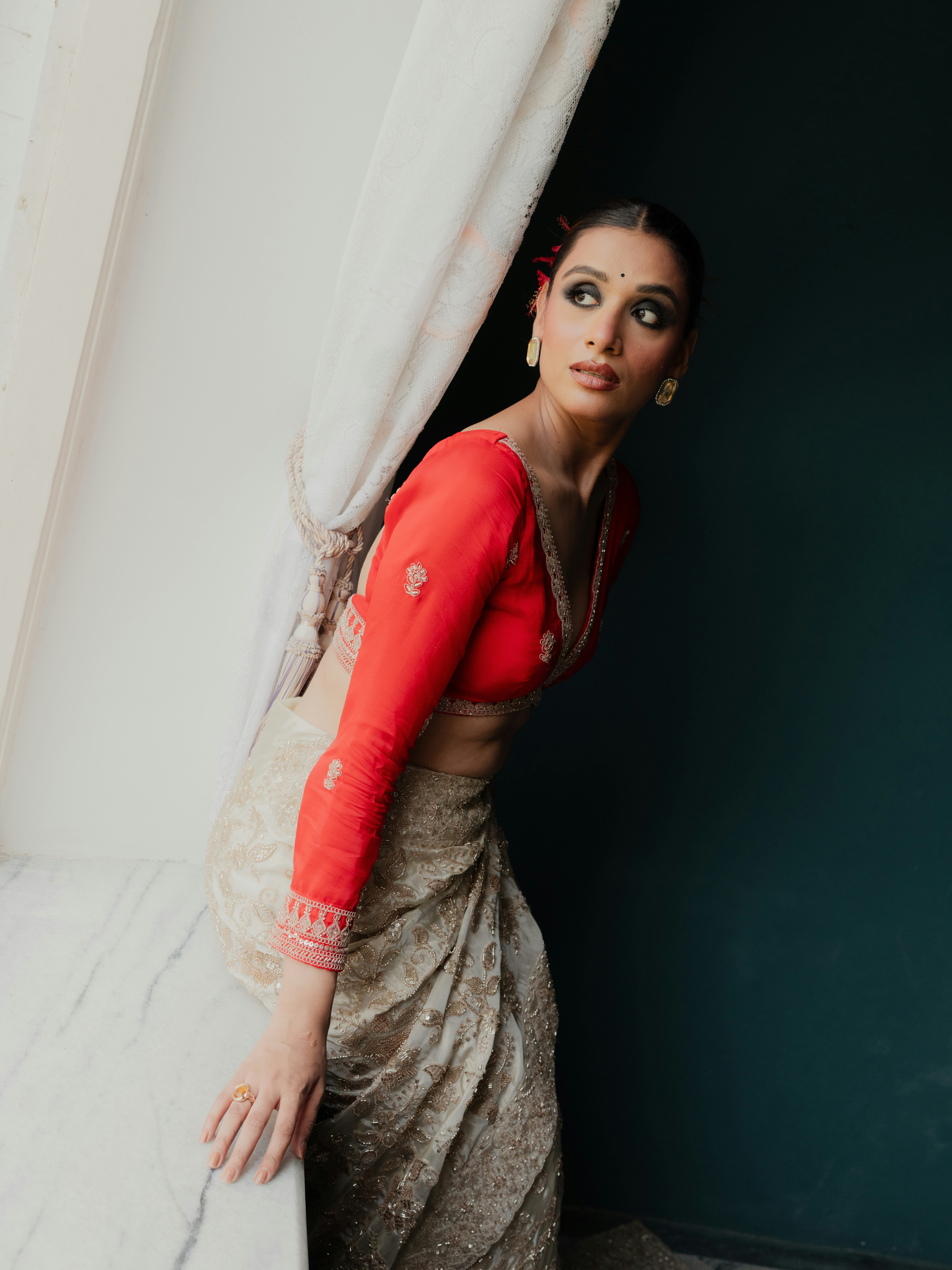 A woman in a red blouse and embroidered cream sari leaning against a curtain