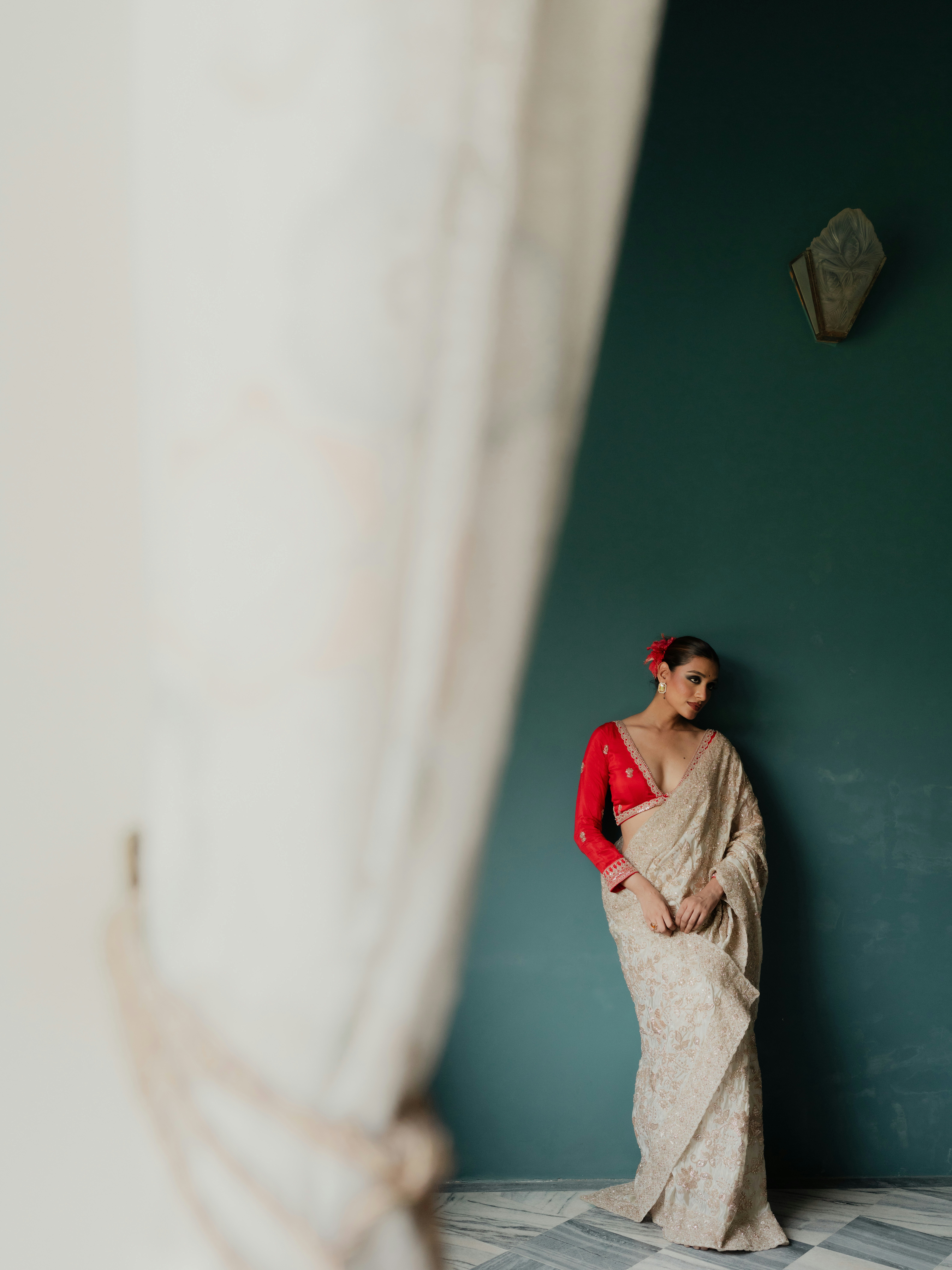 A woman in a red blouse and cream sari against a dark green wall