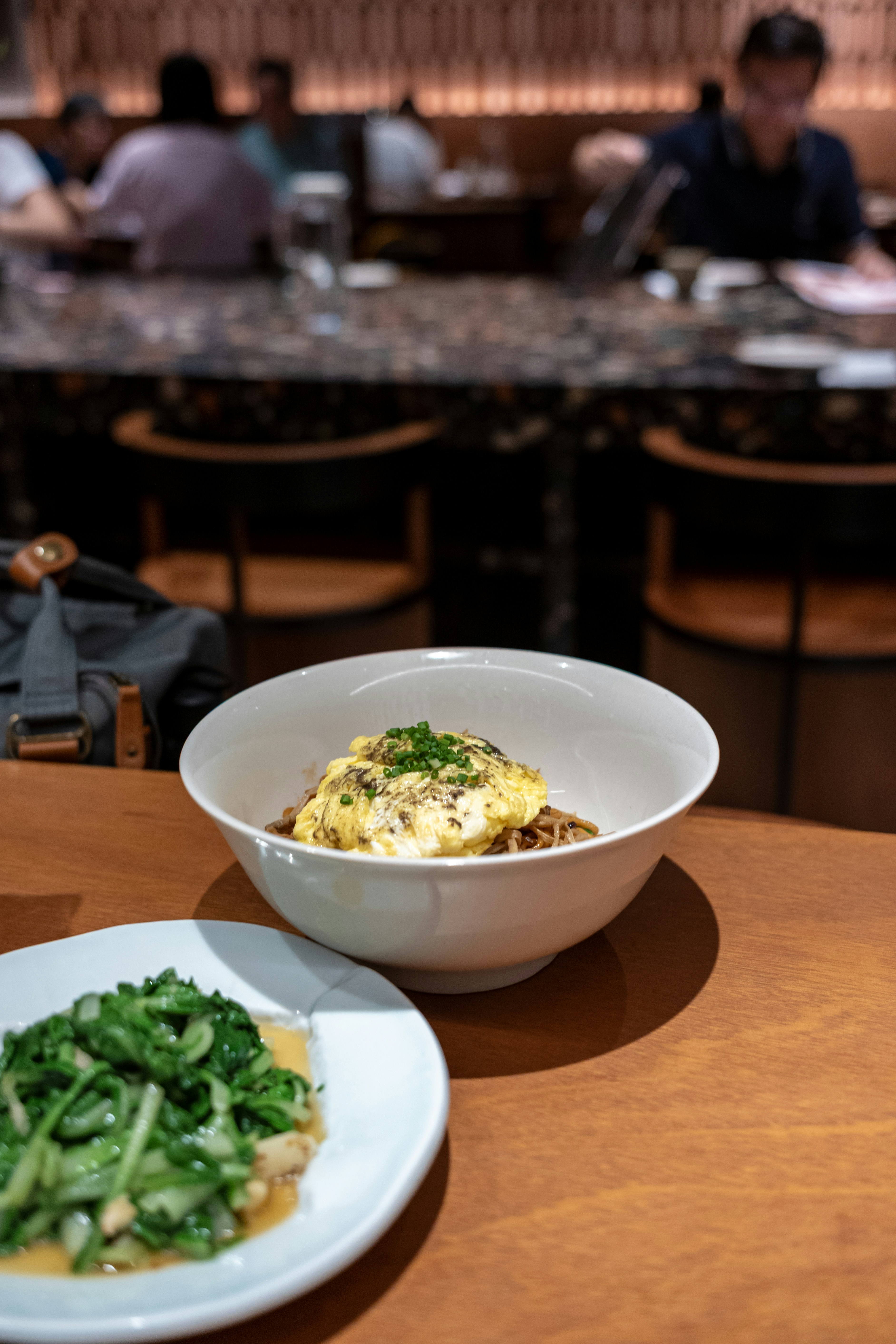 A bowl of noodles topped with egg and chives beside a plate of greens