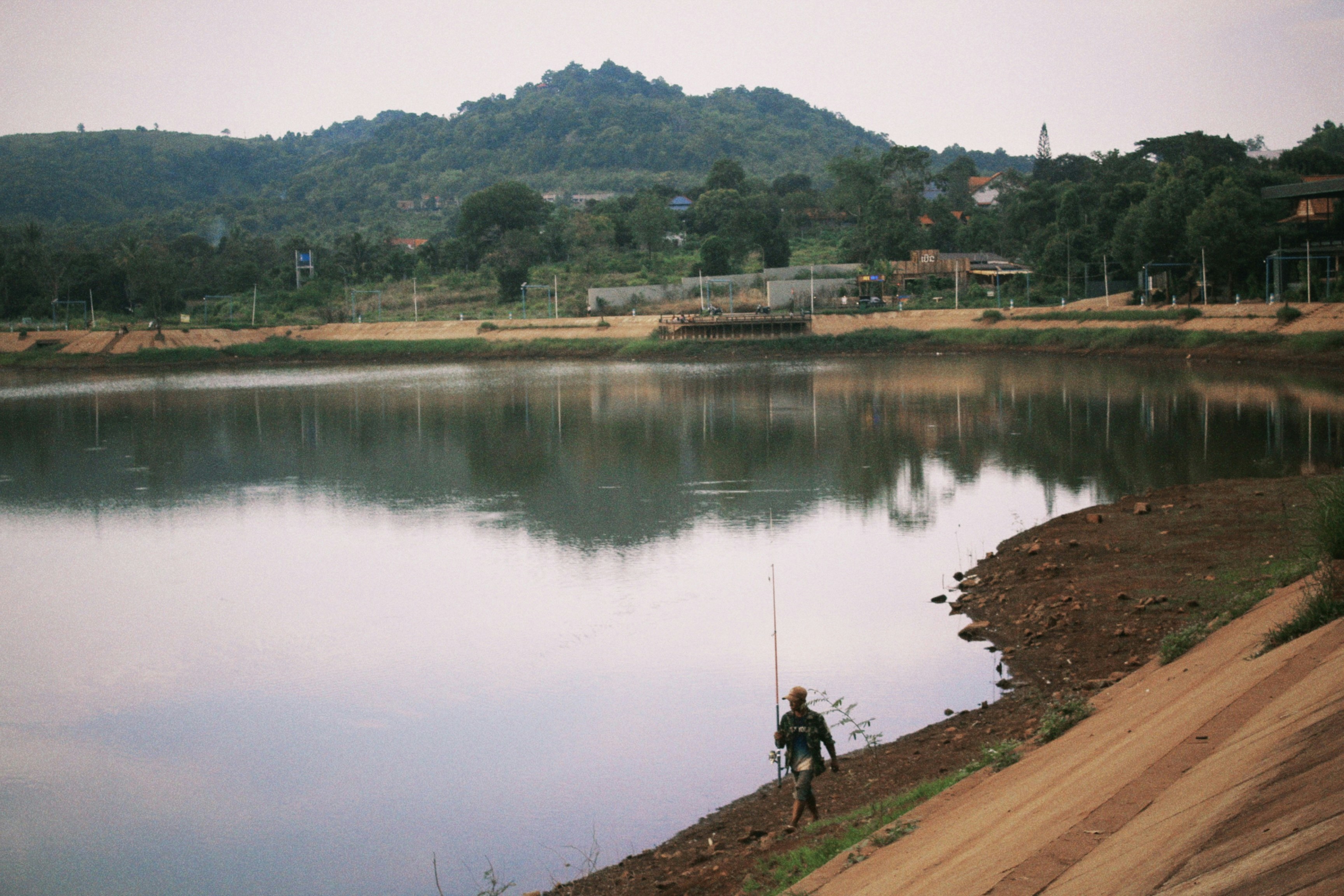A person holding a fishing rod walking along the bank of a lake