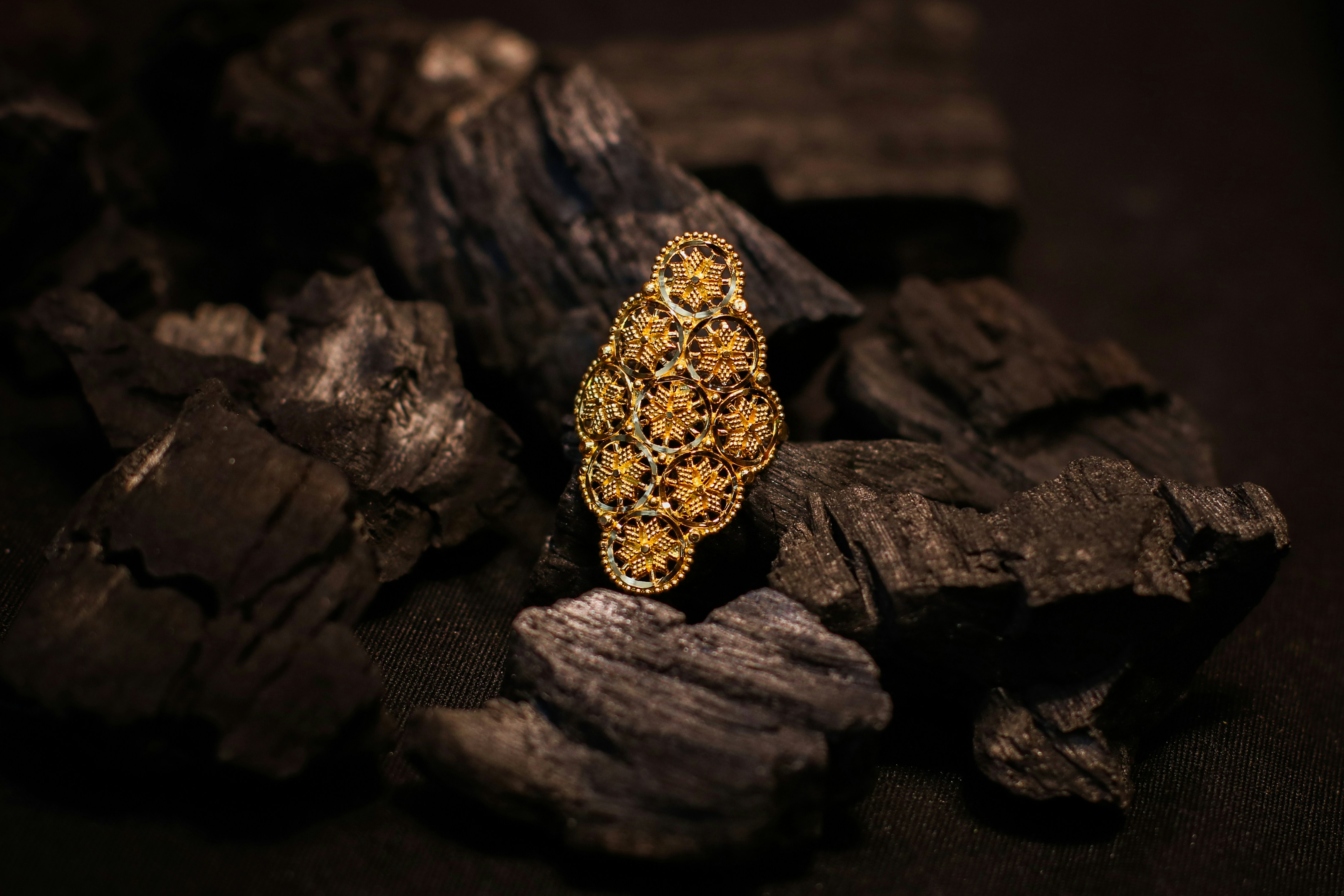 An intricate gold filigree ring resting on a pile of dark charcoal pieces