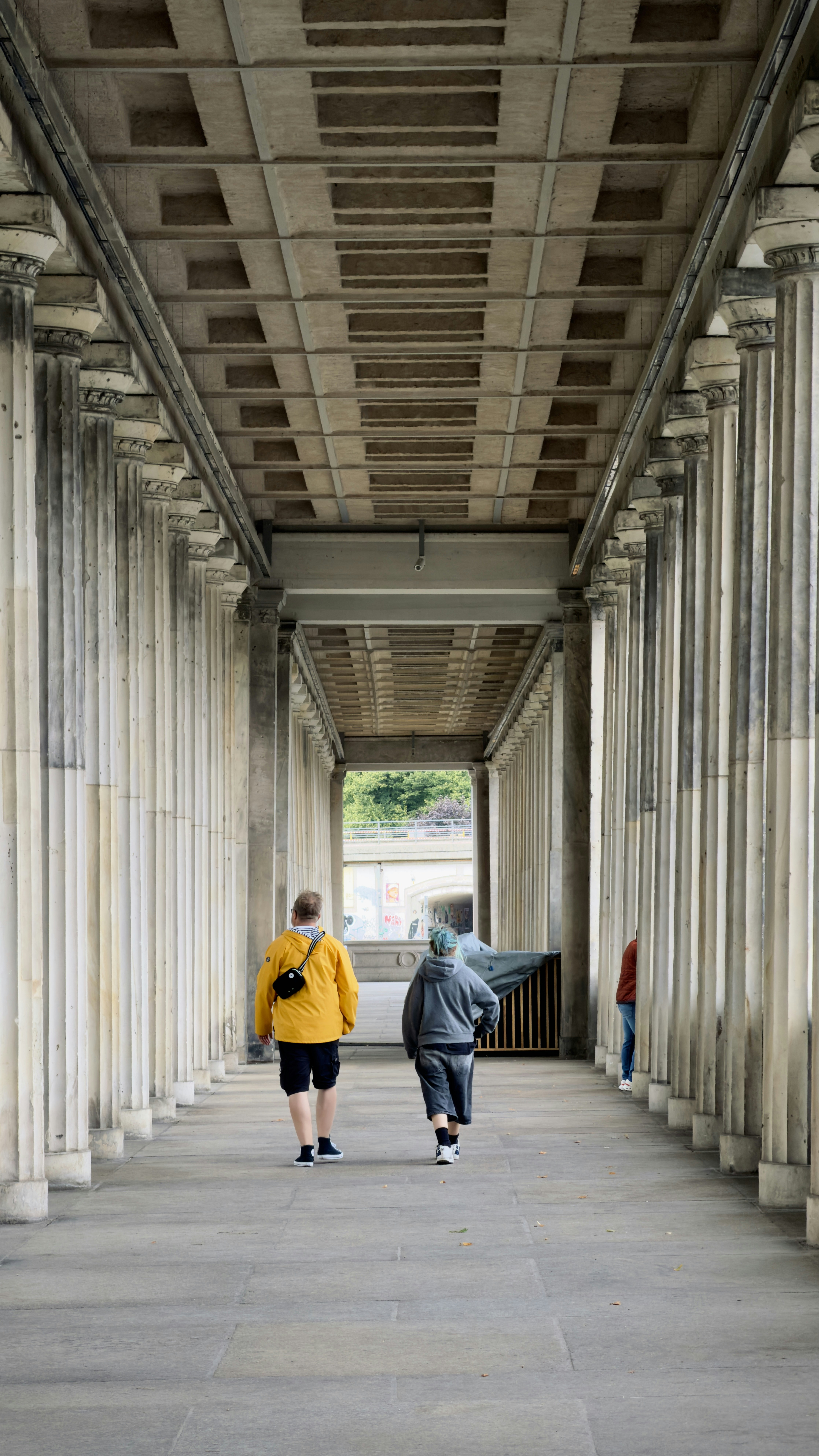 Two people walking through a long colonnade with stone pillars and a coffered ceiling
