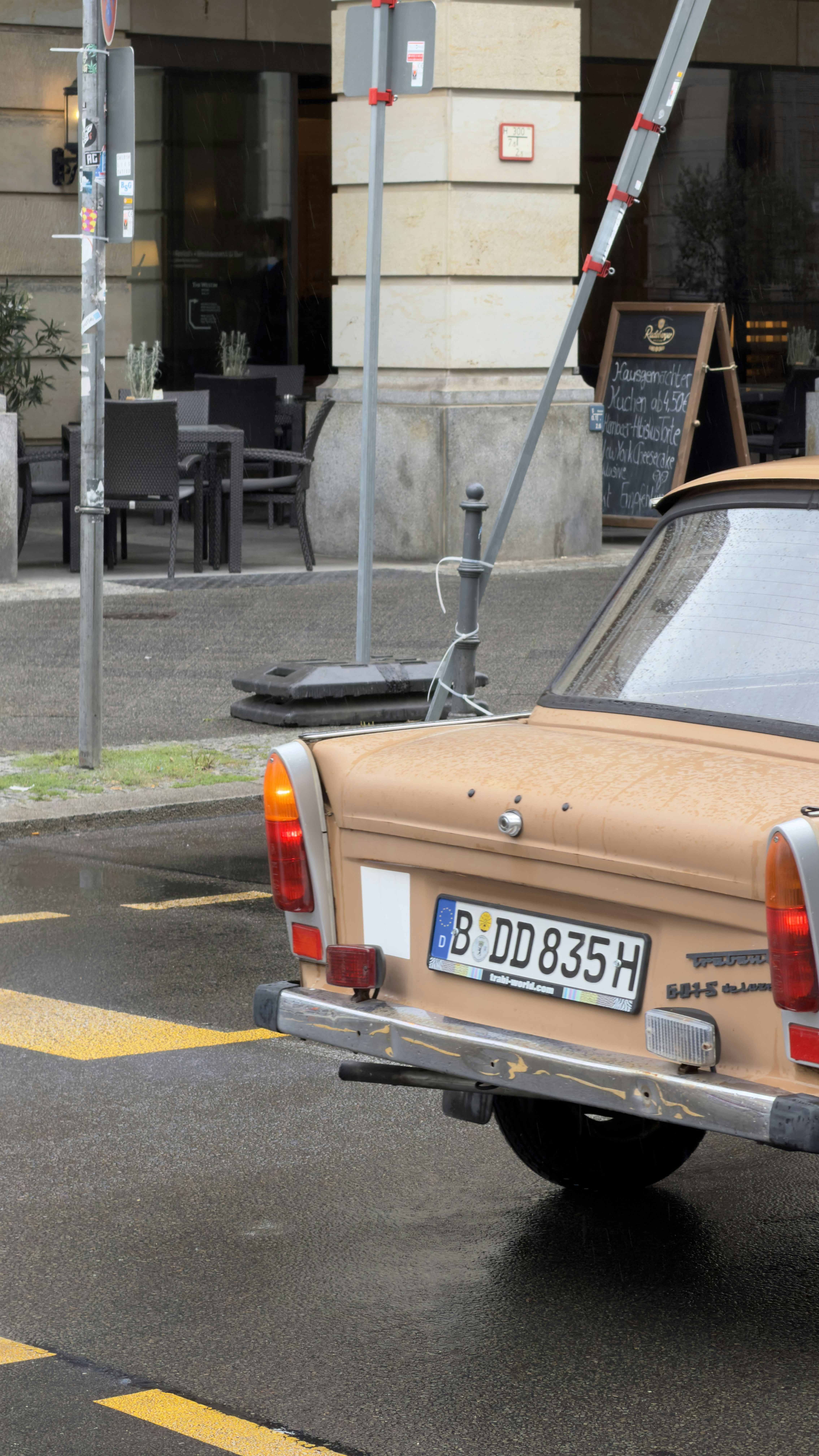 A tan Trabant car parked on a wet city street near a cafe
