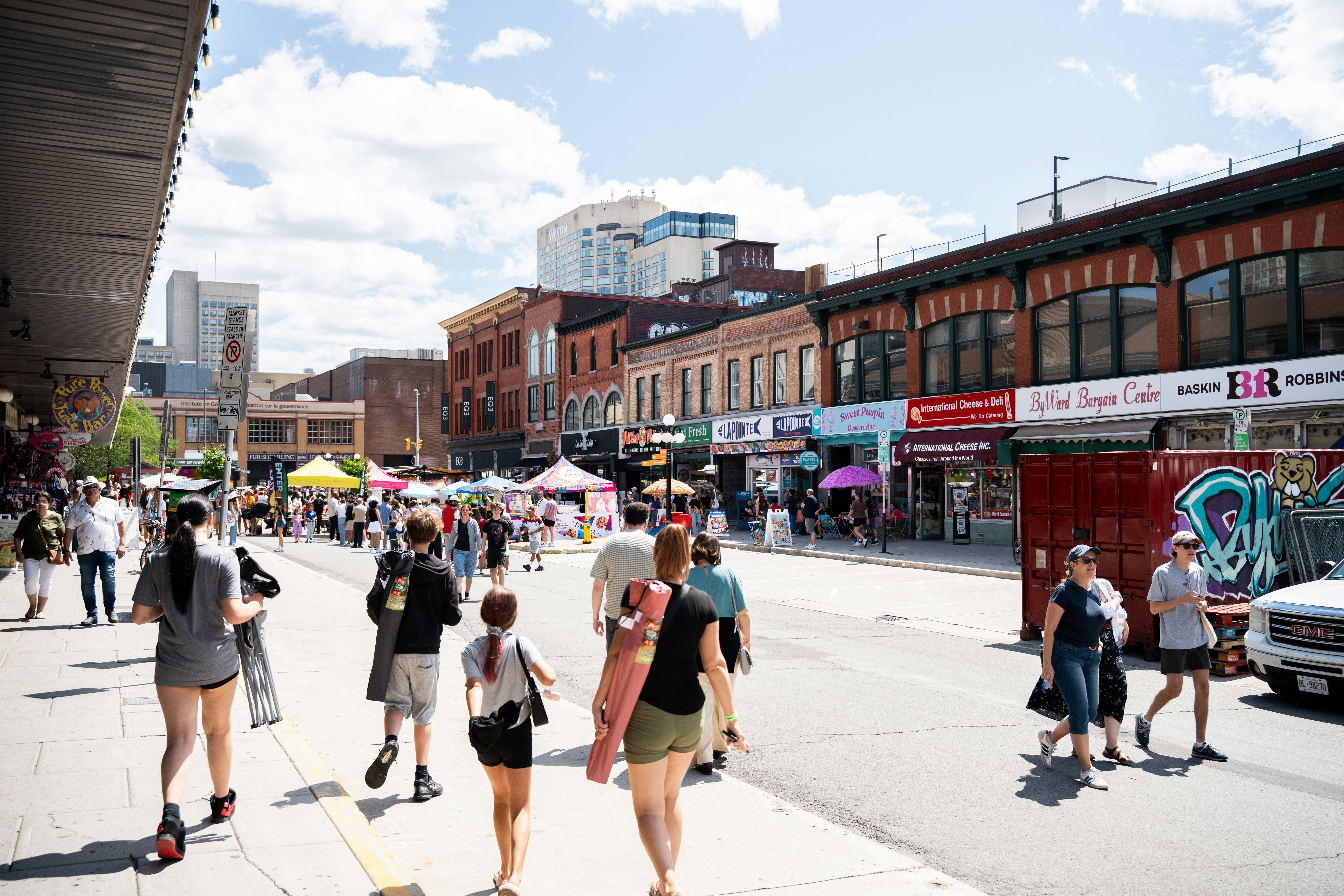 People walking along a sunny street with brick buildings and market stalls in Ottawa