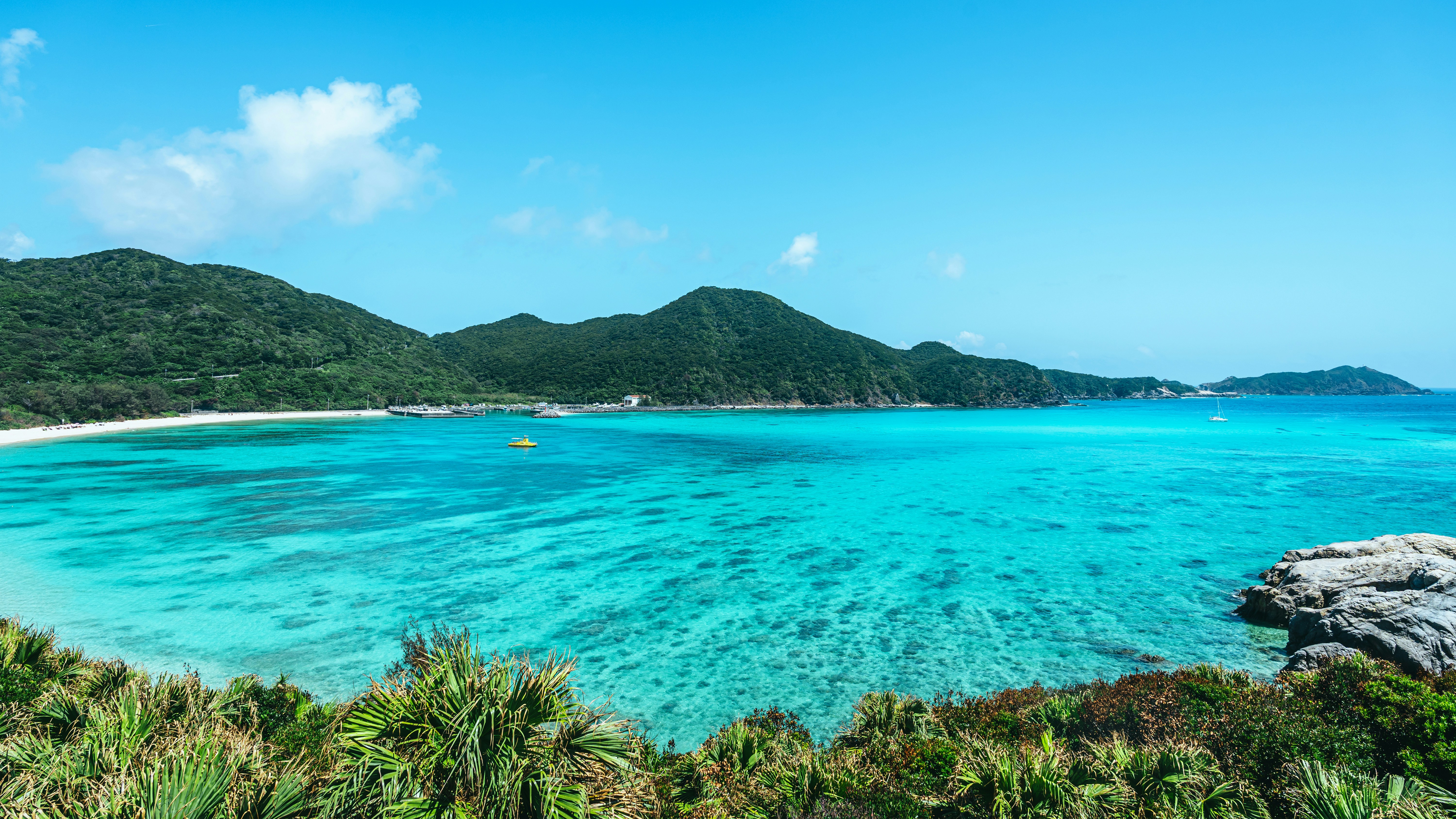 Bright blue water fills a bay on Tokashiki where coral shows dark through the shallows and a pale beach curves under green hills