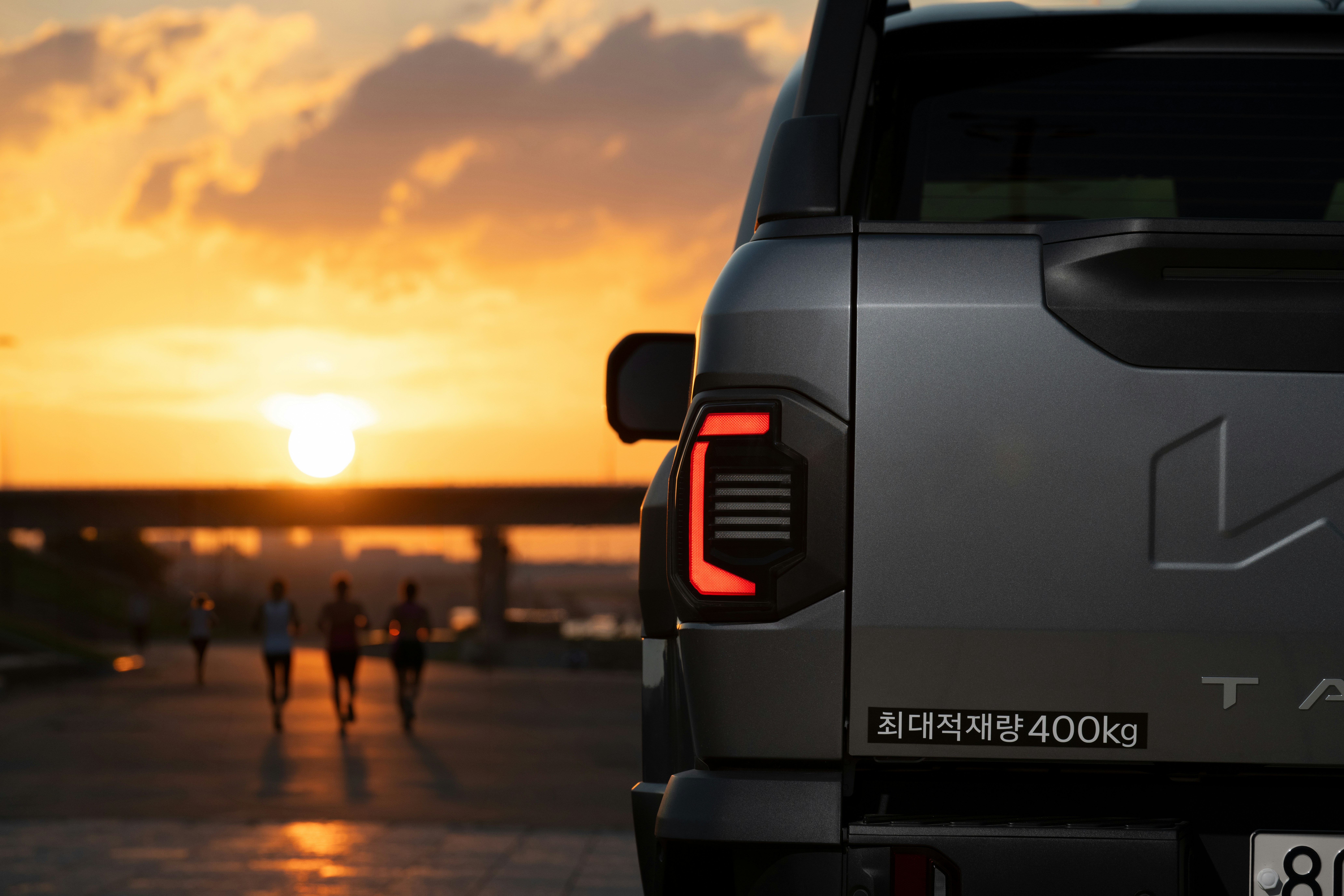 A grey pickup truck tail light with people running on a path at golden hour