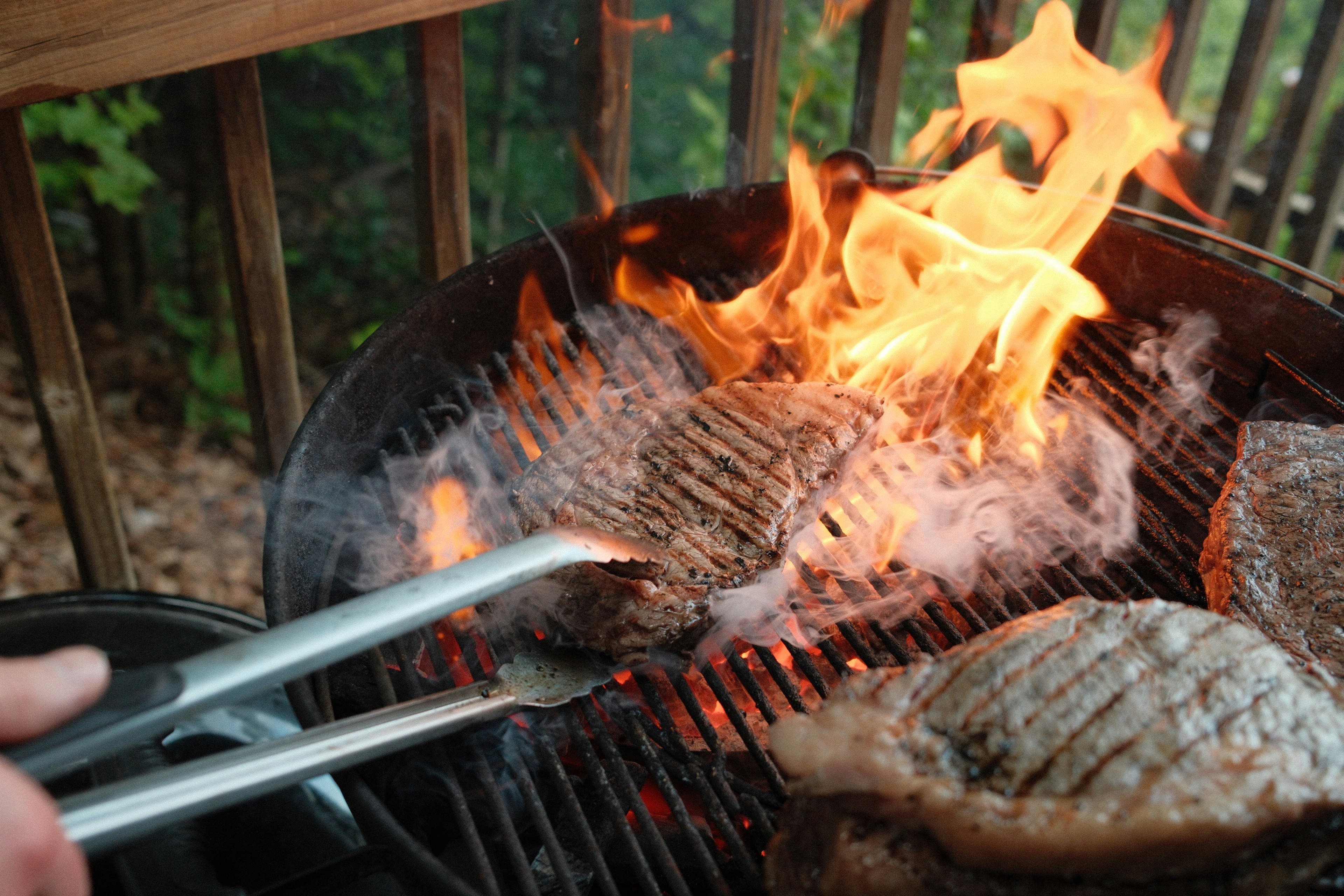 A person using metal tongs to flip a steak on a flaming charcoal grill