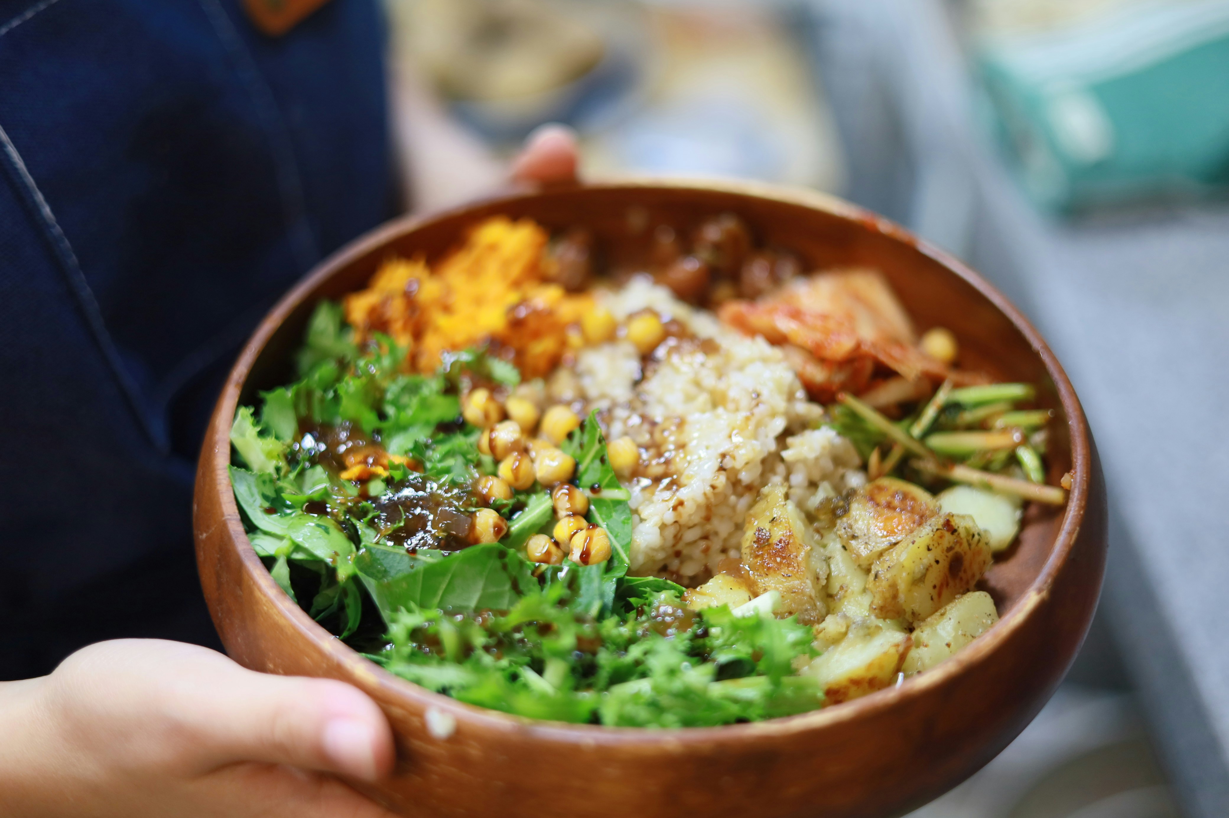 A person holding a wooden bowl filled with rice, chickpeas, greens, and potatoes