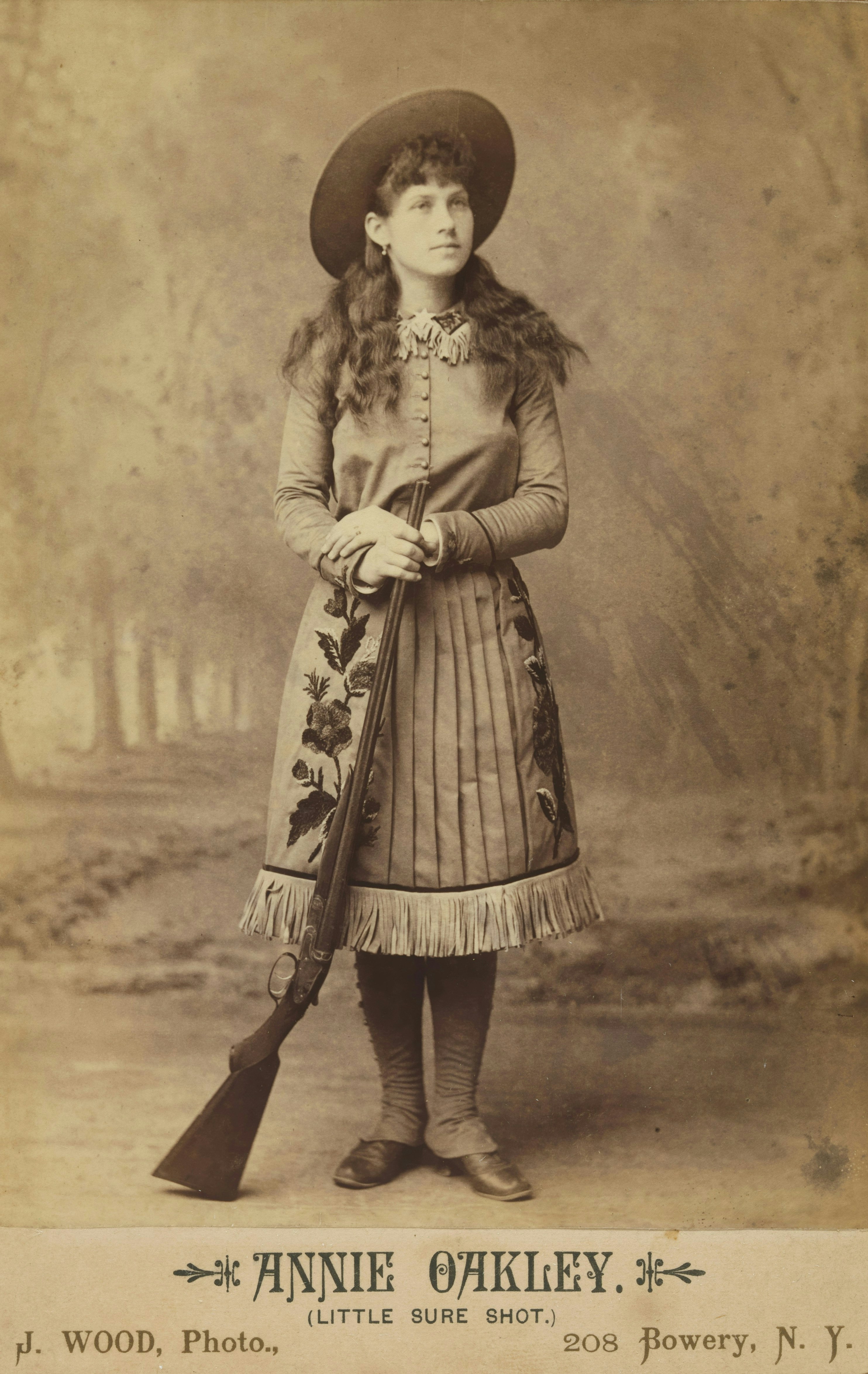 A woman in a wide-brimmed hat and fringed skirt holding a long rifle