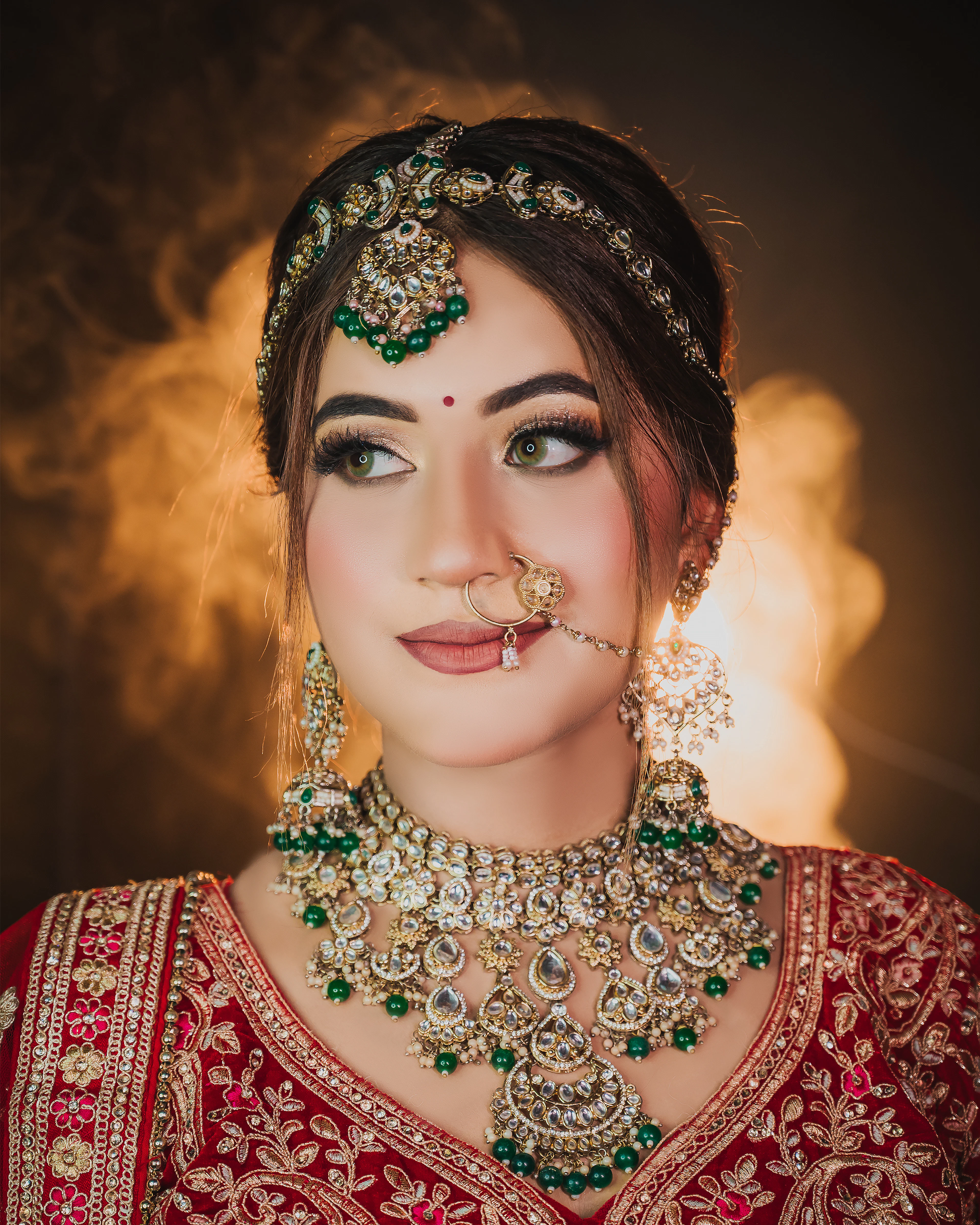 An Indian bride in a red embroidered lehenga wearing a kundan maang tikka, nose ring and an emerald-and-gold necklace