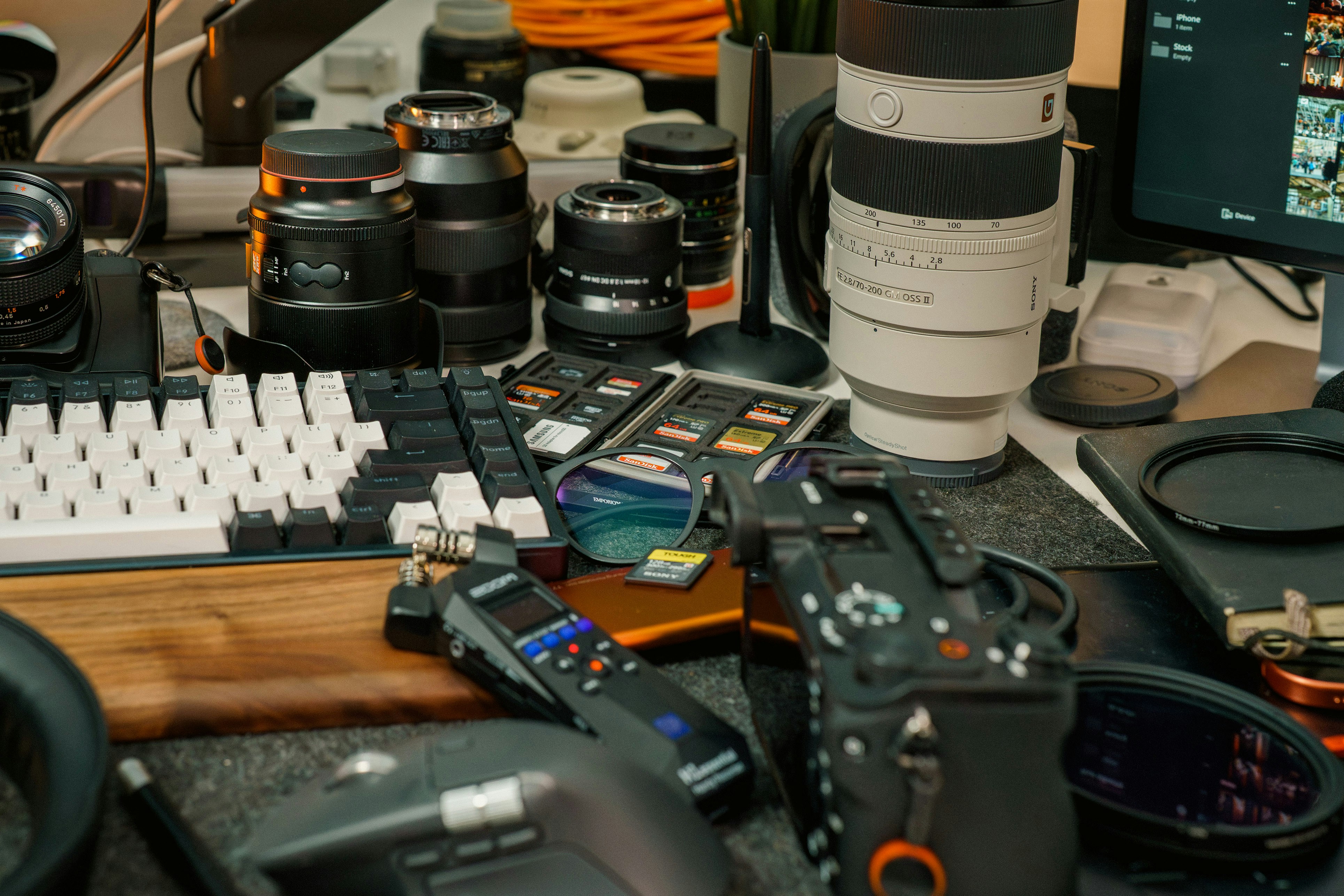 Camera lenses, a keyboard, and audio equipment on a cluttered desk workspace
