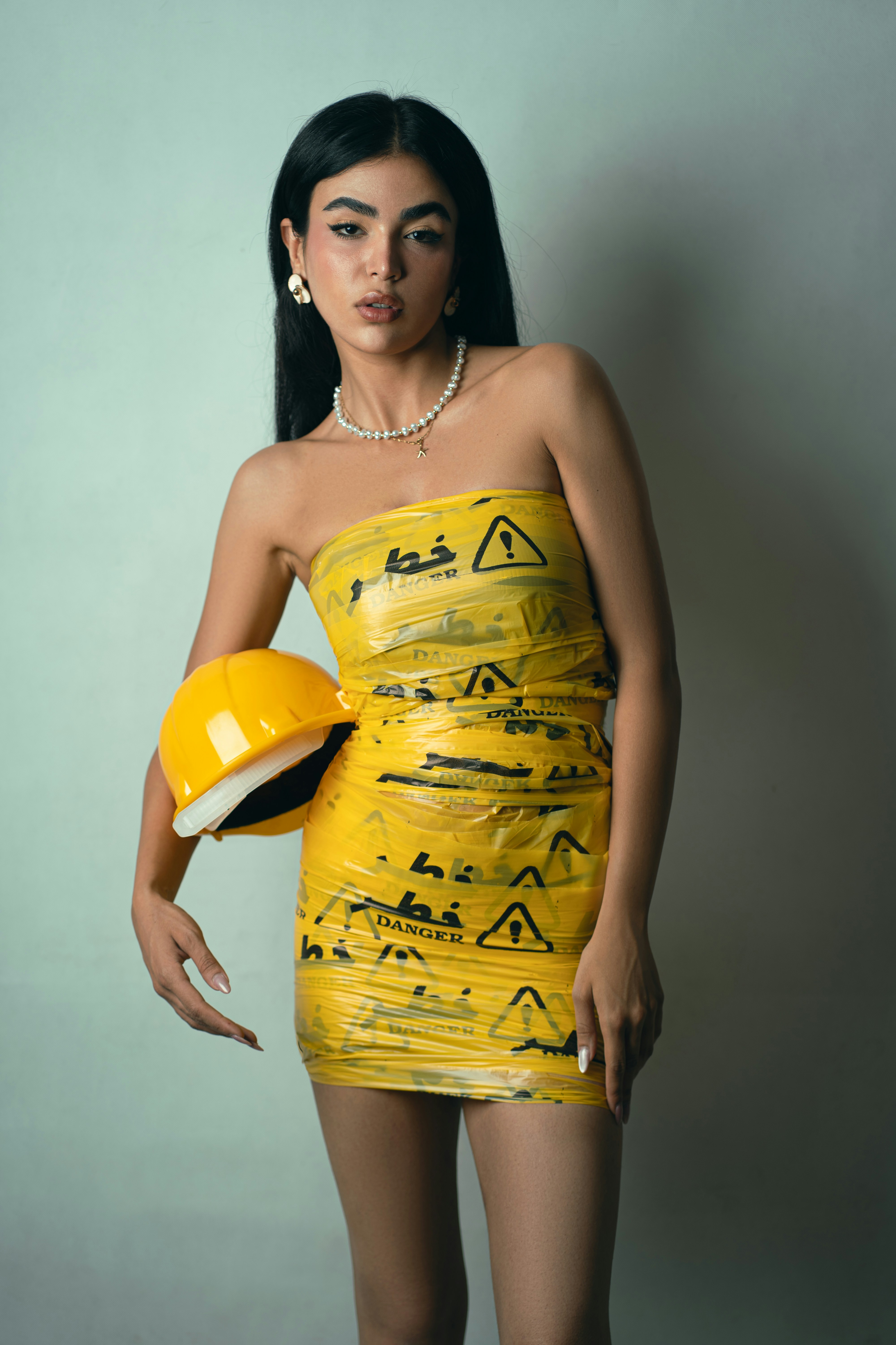 A woman wearing a dress made of yellow danger tape holding a hard hat