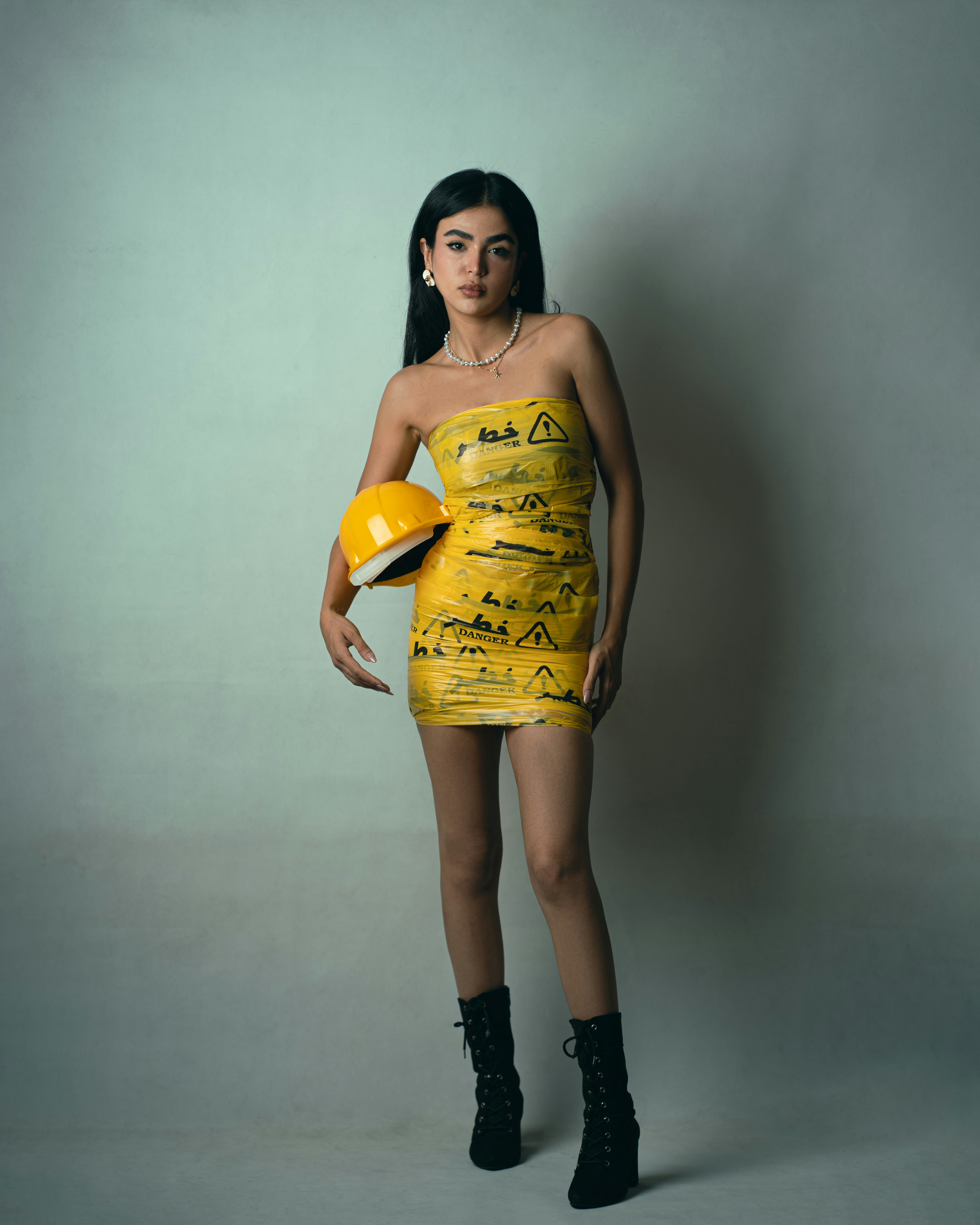 A woman wearing a dress made of yellow danger tape holding a hard hat