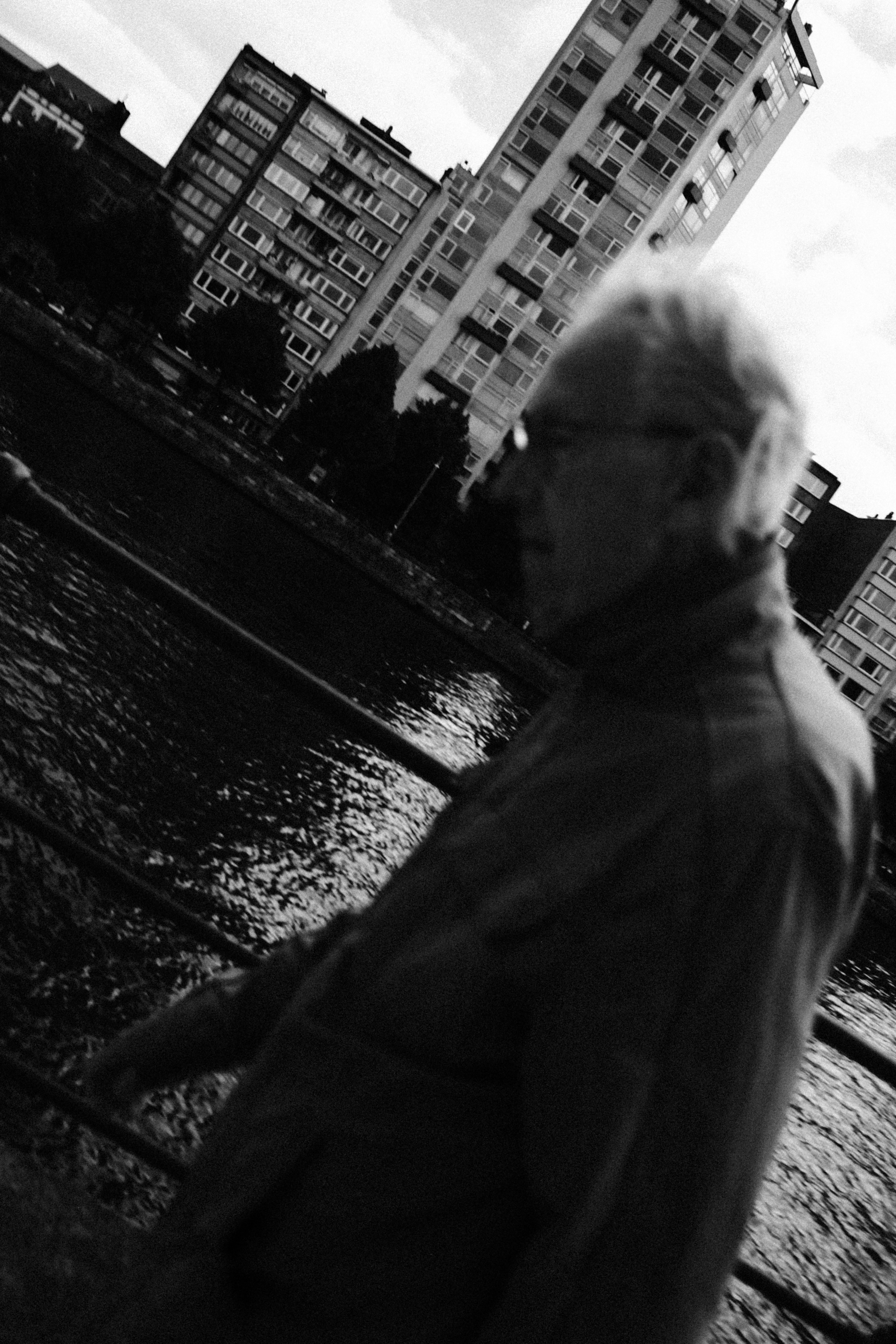 A person in glasses walking along a riverbank with tall apartment buildings behind