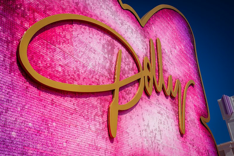 Gold cursive Dolly signature mounted on a giant hot-pink sequined heart, shot from below against a deep blue sky