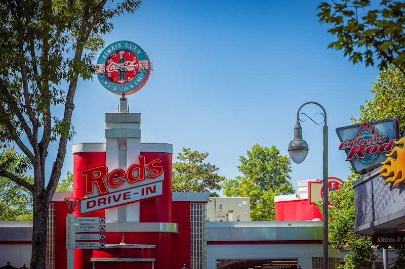 Red-and-white 1950s-style drive-in building topped by a round Coca-Cola clock, with a neon Lightning Rod coaster sign at the right edge under a clear blue sky