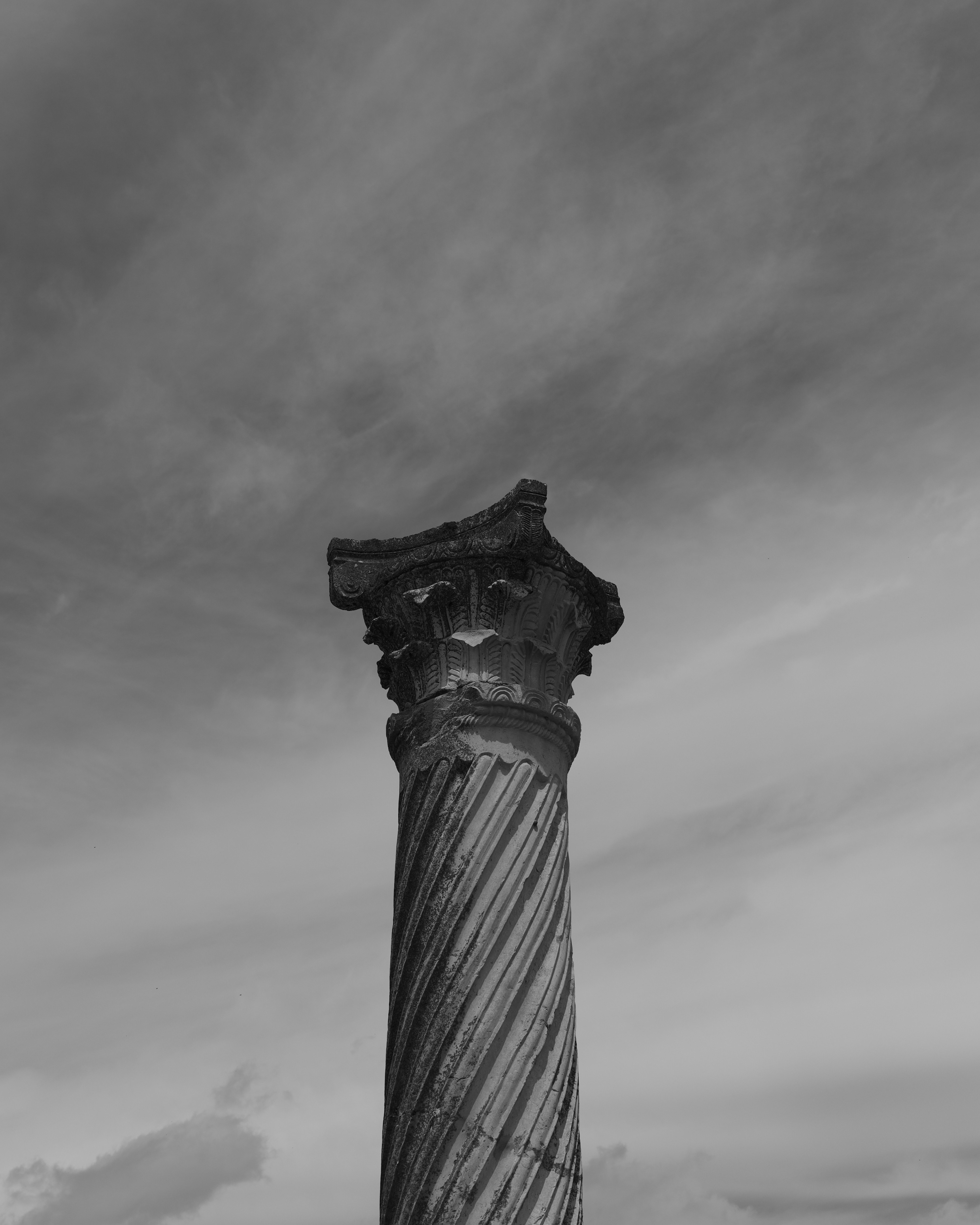 A weathered stone column with a spiral fluted shaft against a cloudy sky