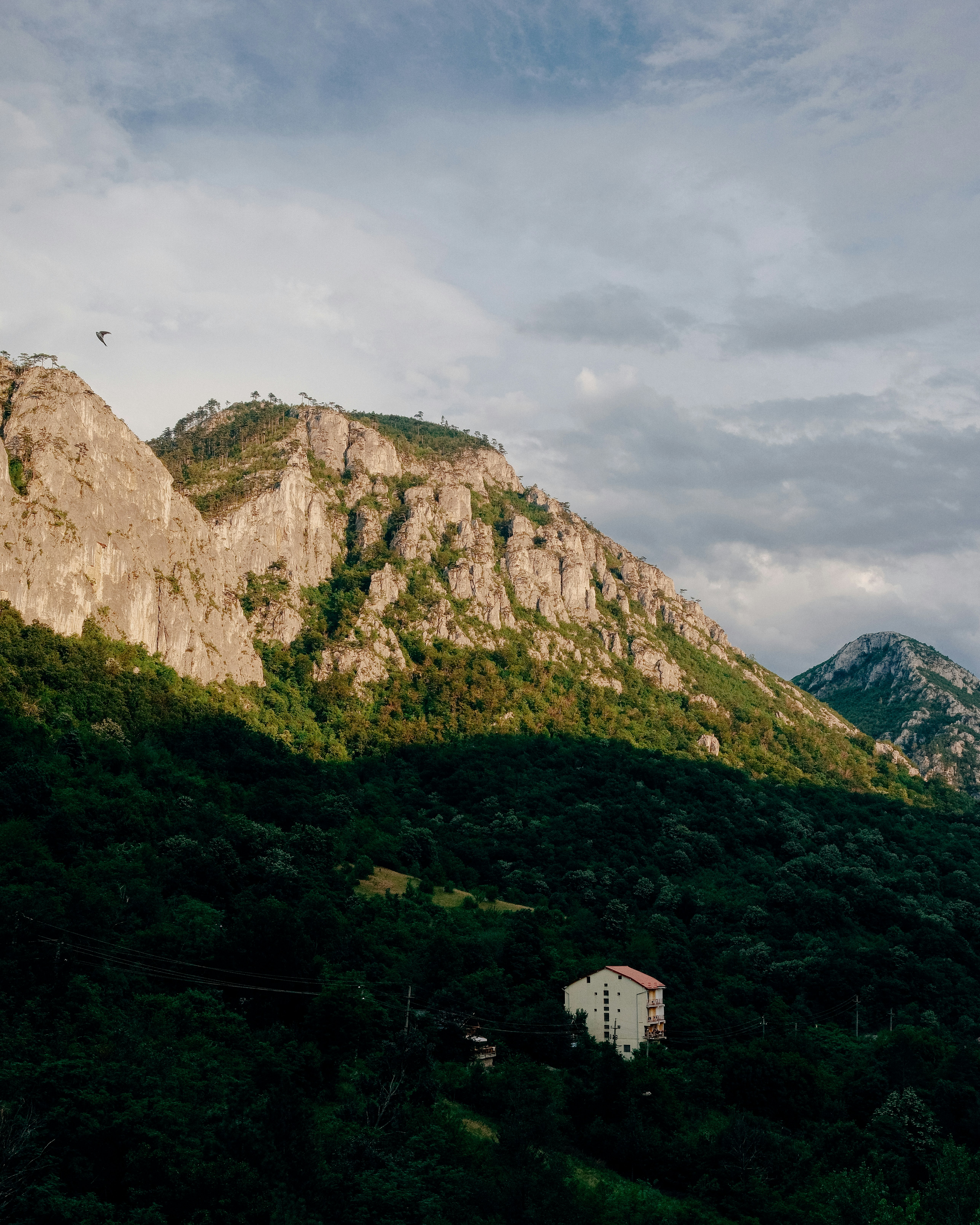 A small house nestled in a lush green forest below towering rocky mountain cliffs