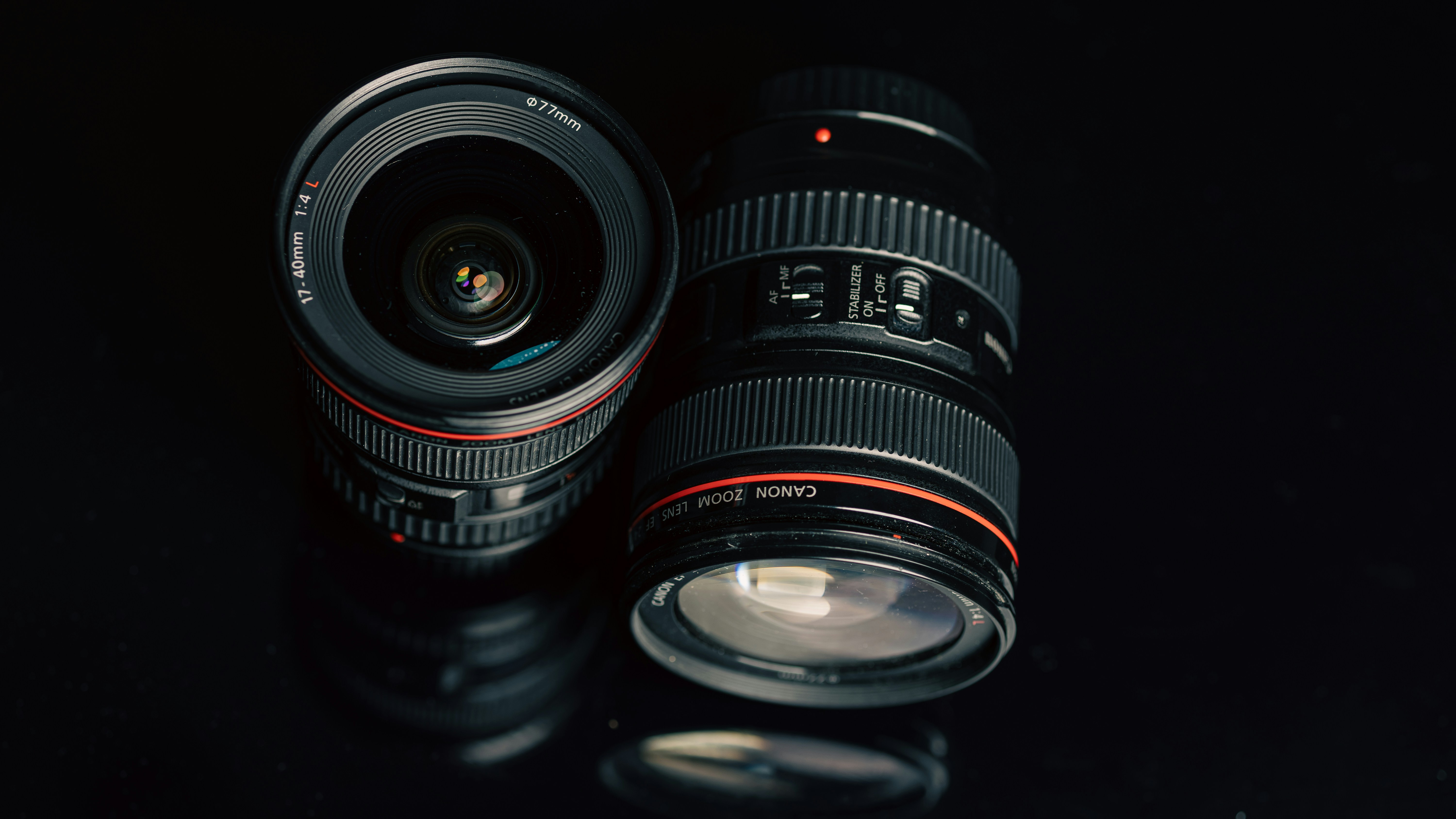 Two black camera lenses with red rings on a reflective black surface