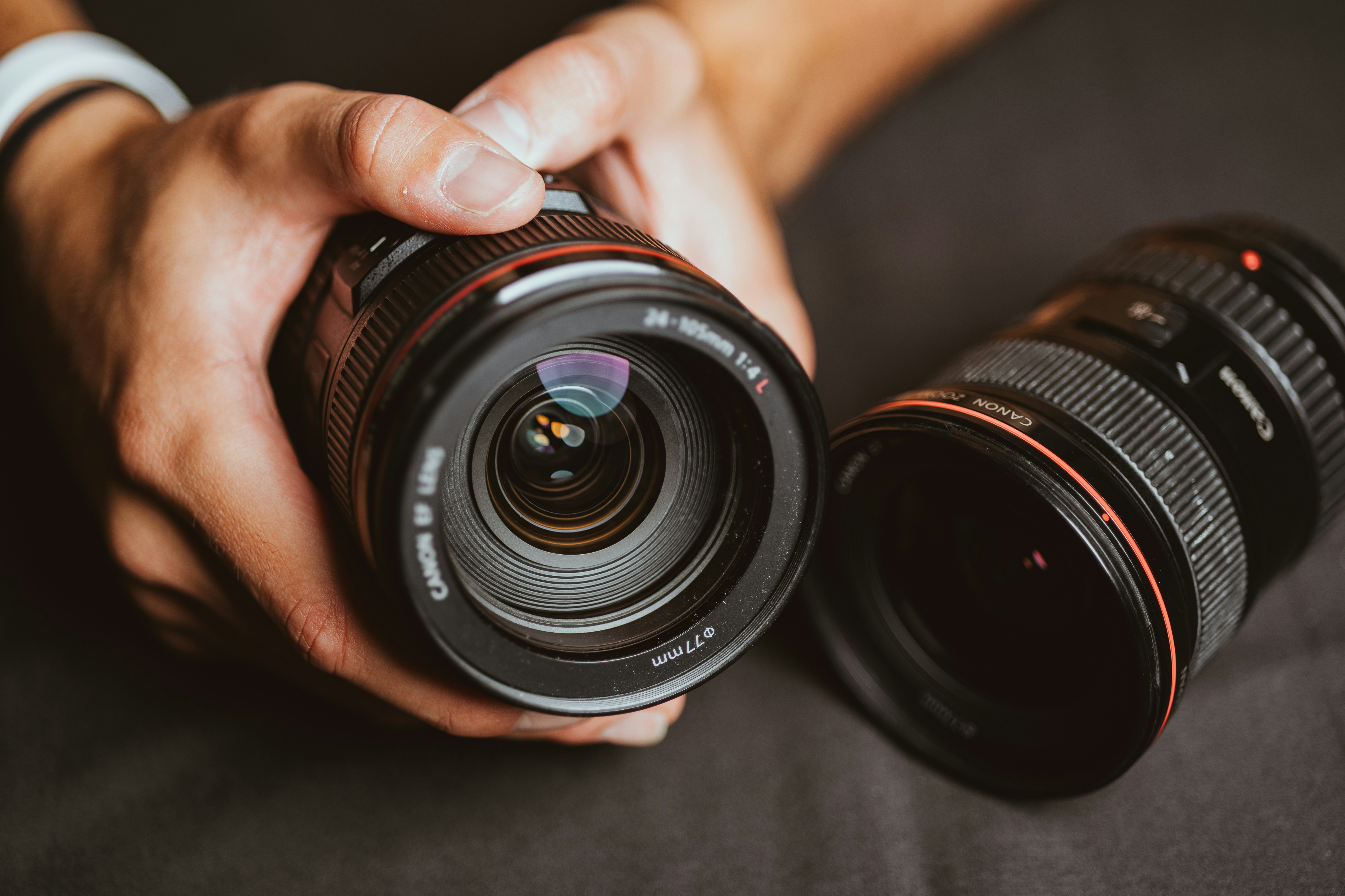 Hands holding a camera lens beside another lens on a dark surface