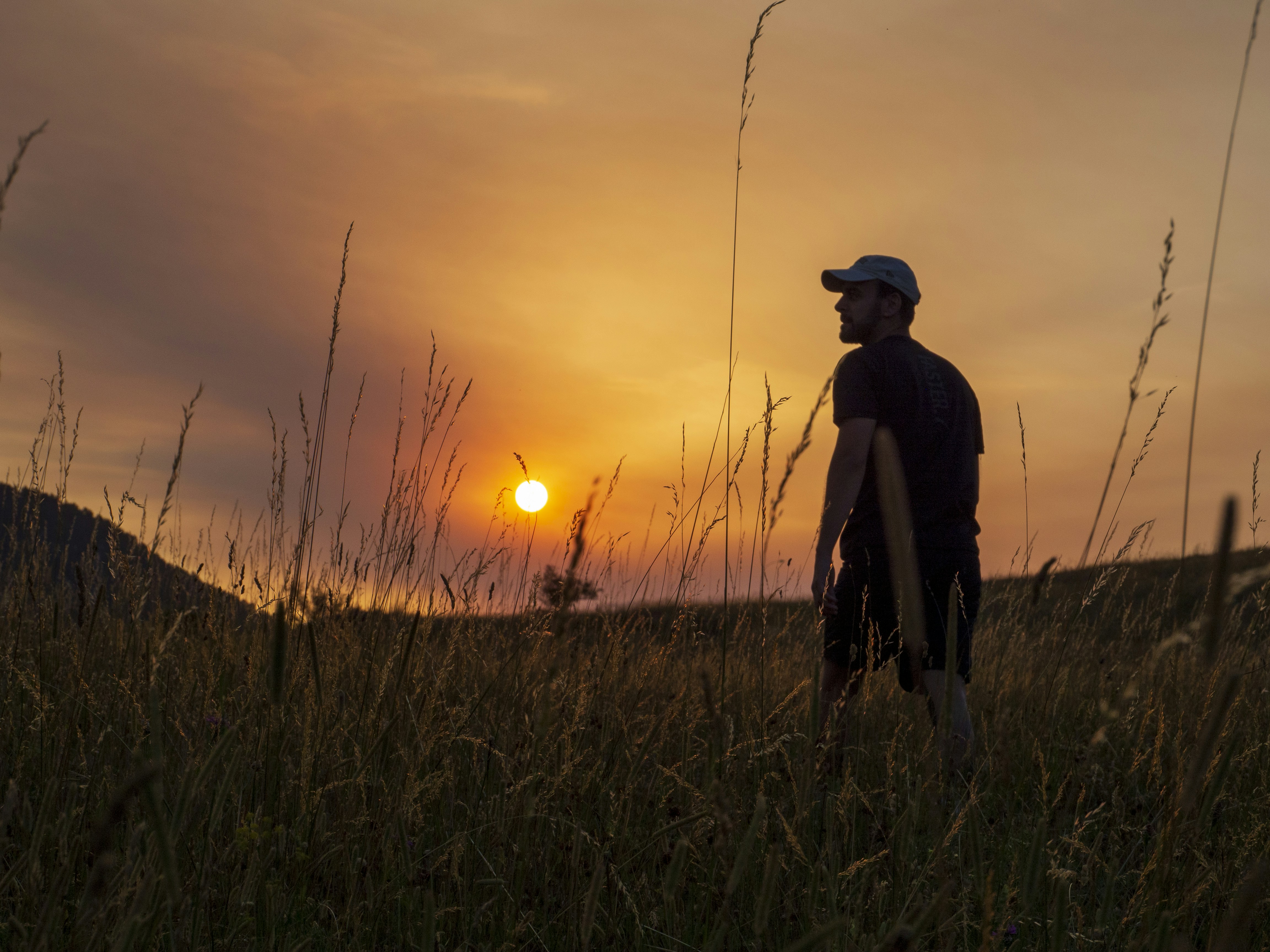 Une personne en casquette marchant à travers de hautes herbes sous un ciel orange chaud