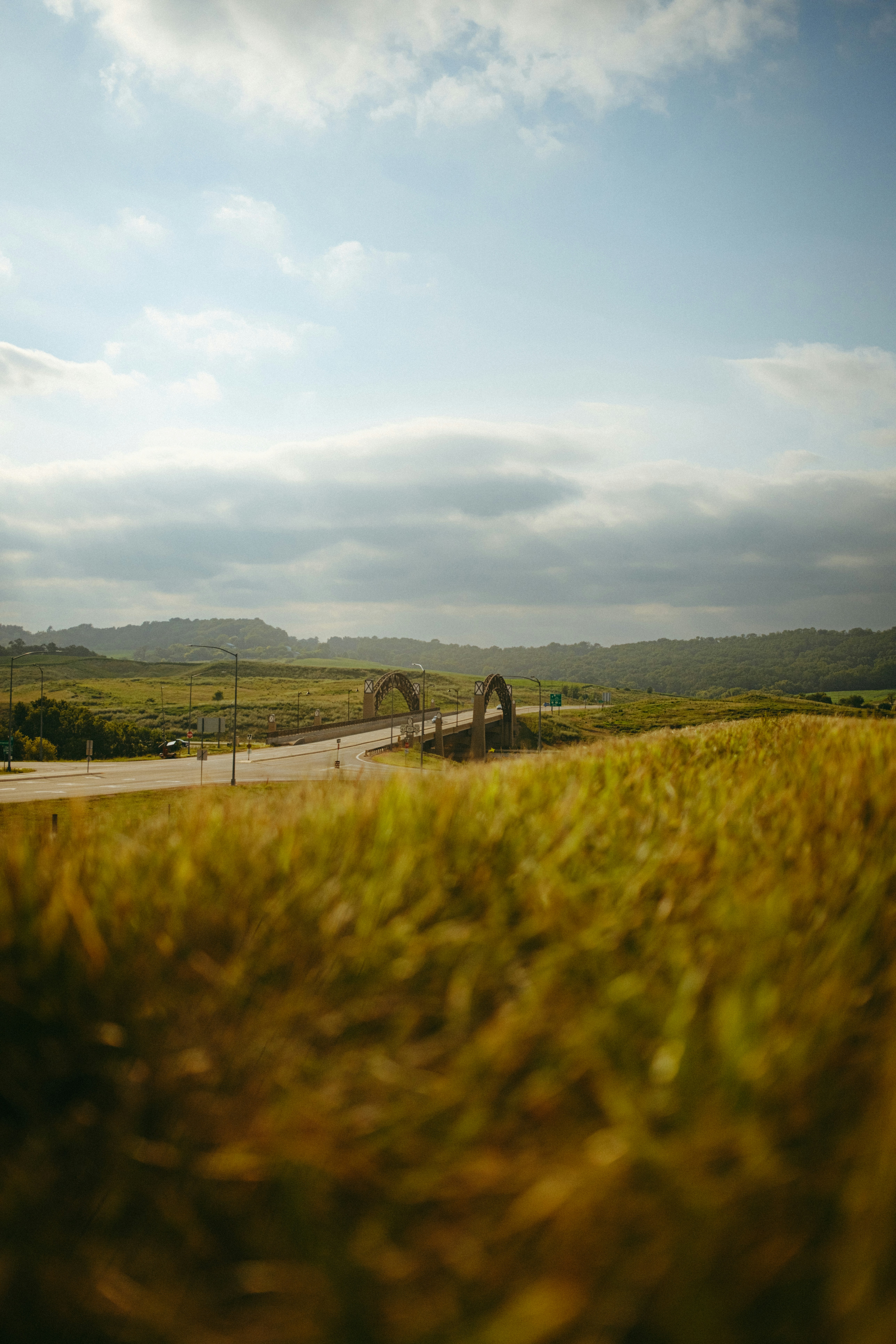 A road crossing an arched bridge through a green hilly landscape under clouds