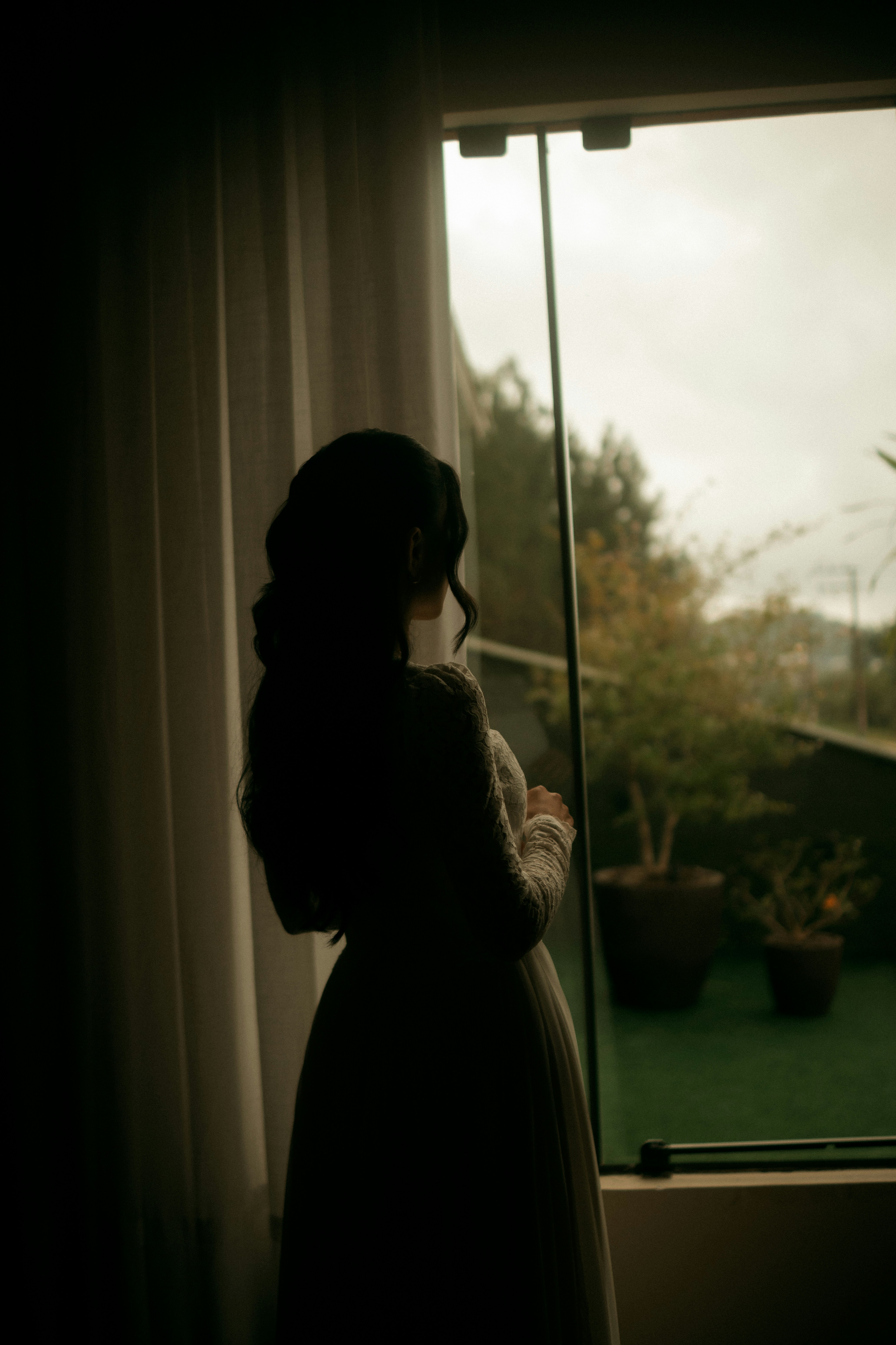 Une femme en silhouette contre une fenêtre avec des rideaux transparents et des plantes en pot