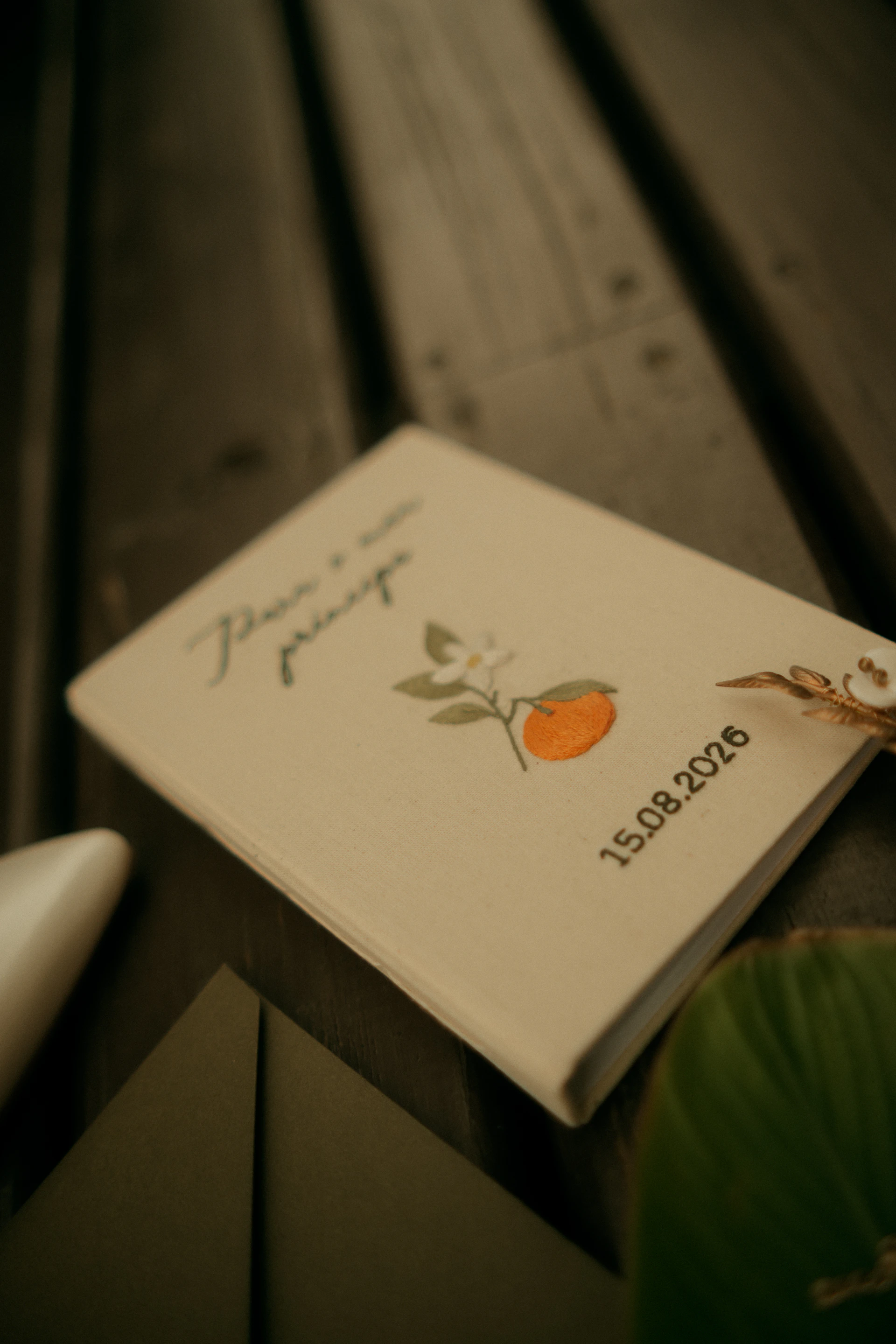 A cream-colored notebook with embroidered orange fruit and leaves on a dark wooden surface