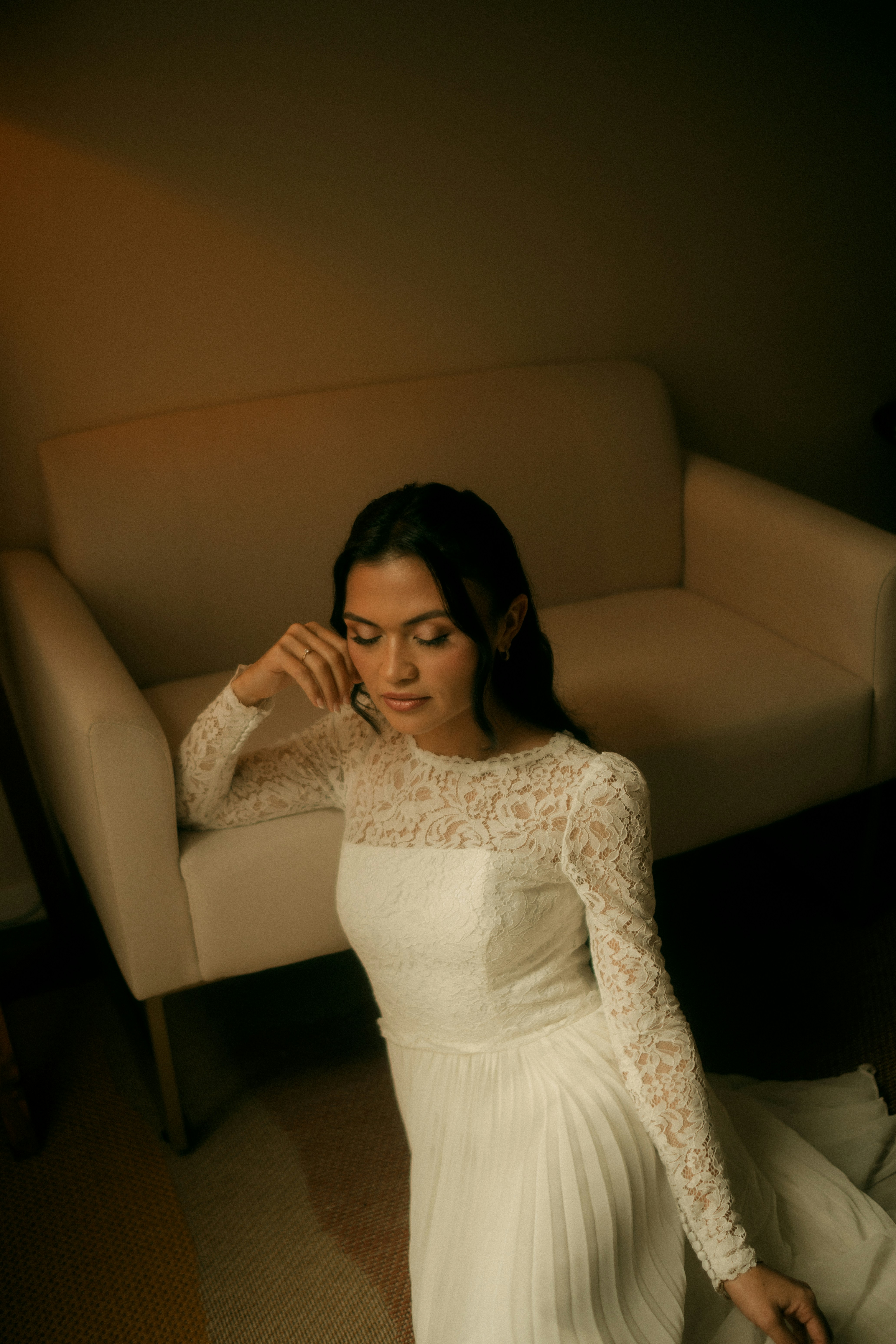 Une femme en robe de mariée en dentelle blanche assise devant un canapé beige