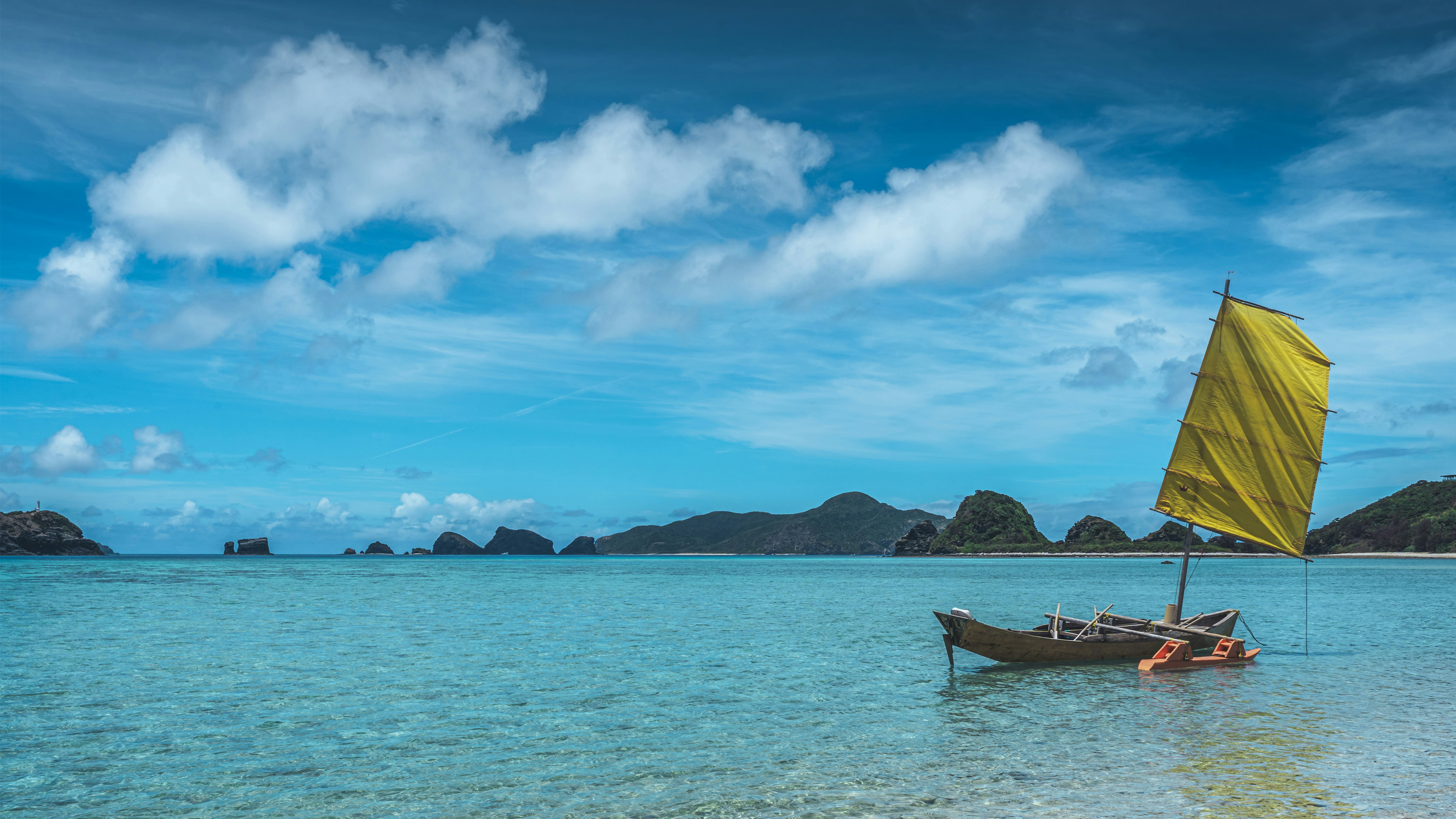 A wooden sabani boat with a yellow sail floats in clear shallow water off Zamami while the jagged outline of smaller Kerama islands lines the horizon