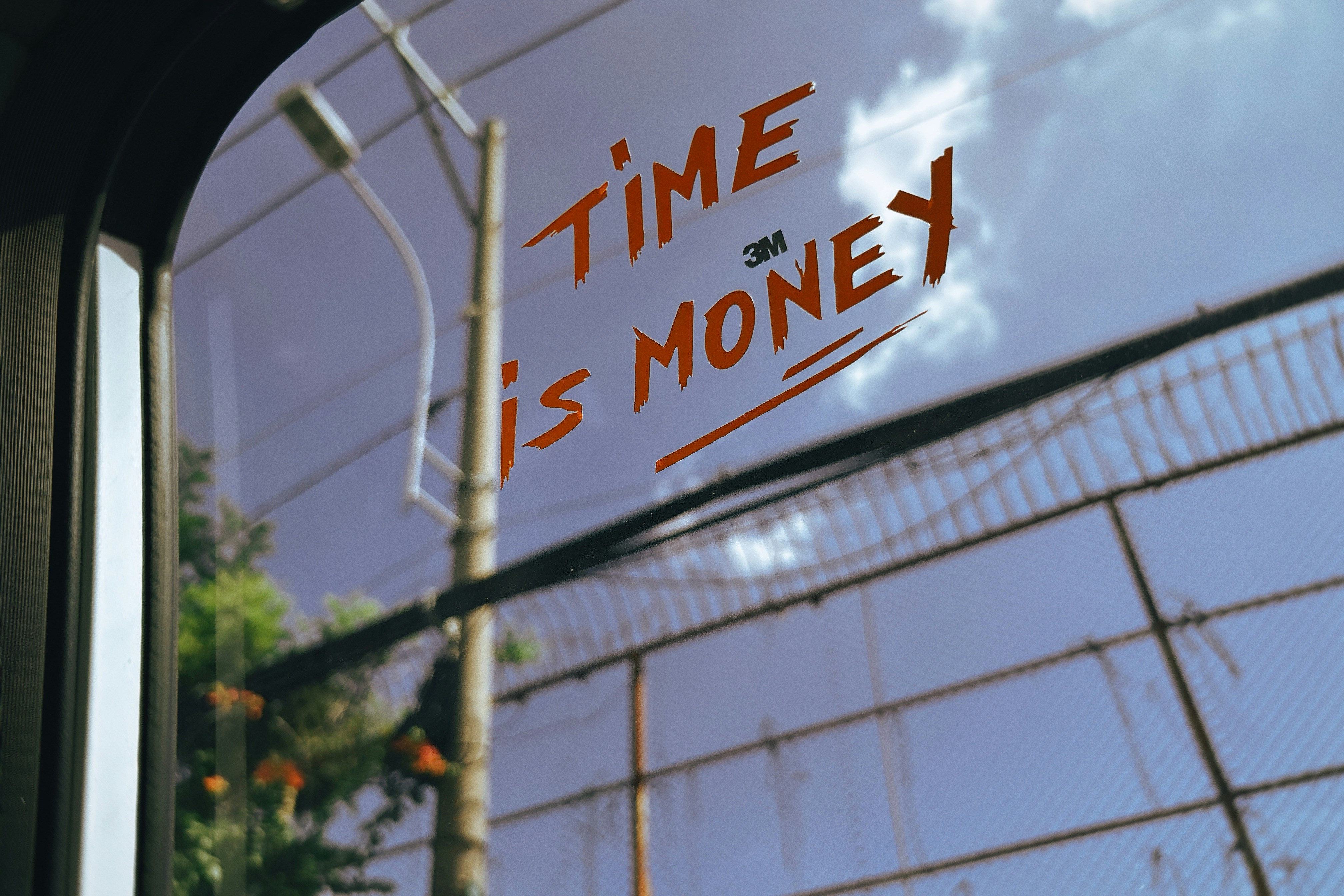 Orange text reading "TIME IS MONEY" on a car window against a chain-link fence