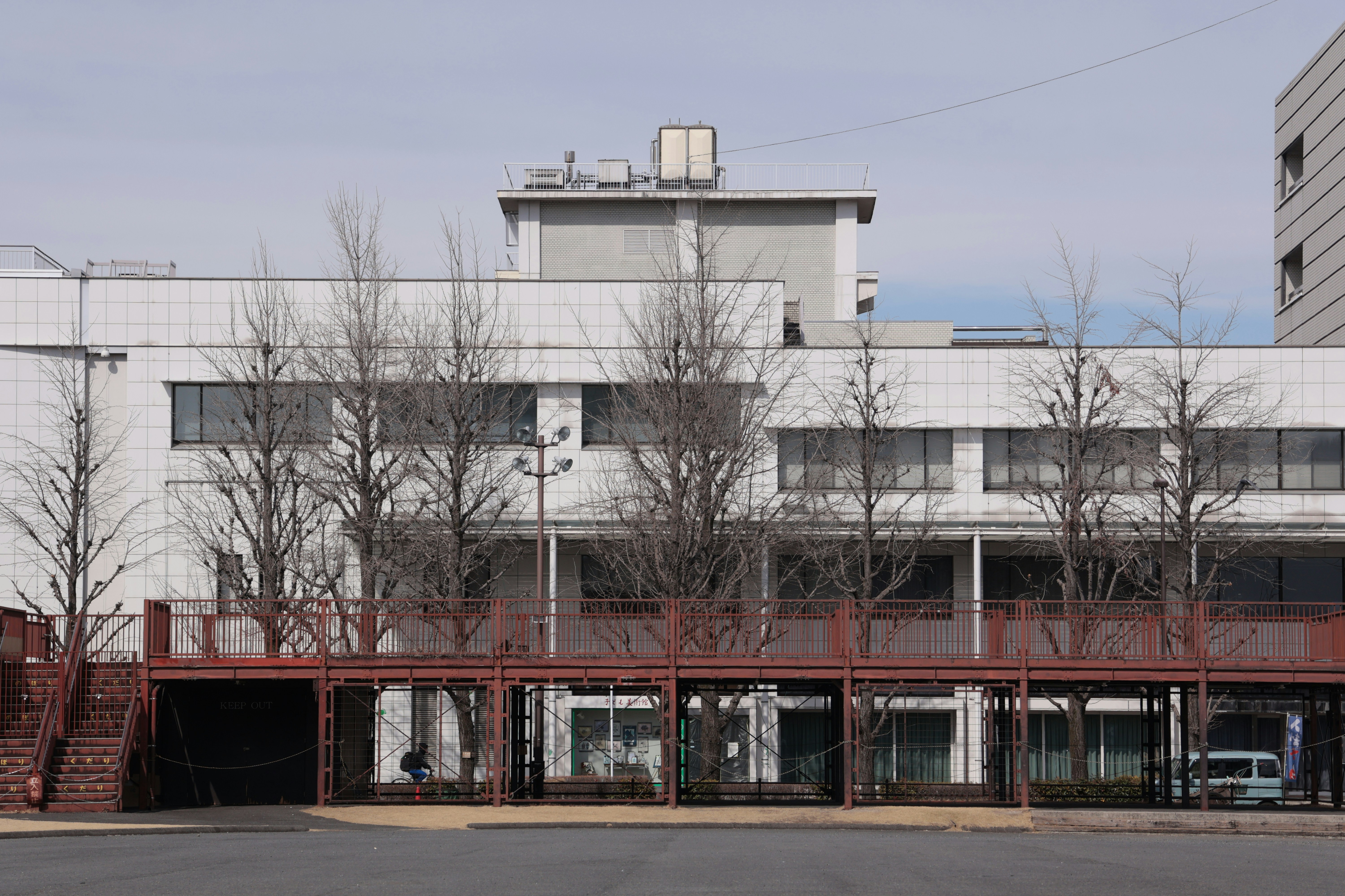 赤い金属製歩行者橋の背後に白いタイル張りの建物と裸の木々がある