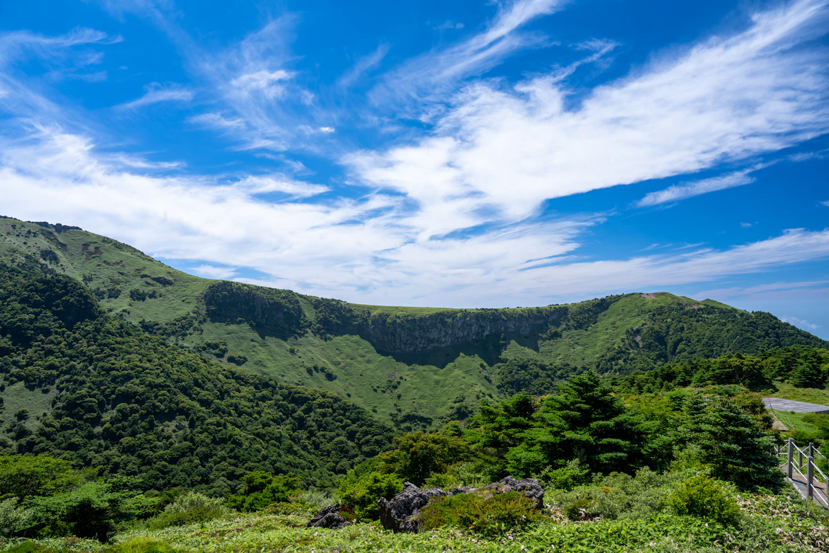 Green volcanic slopes and cliffs under a blue sky near Seogwipo, Jeju, with a wooden railing at the right
