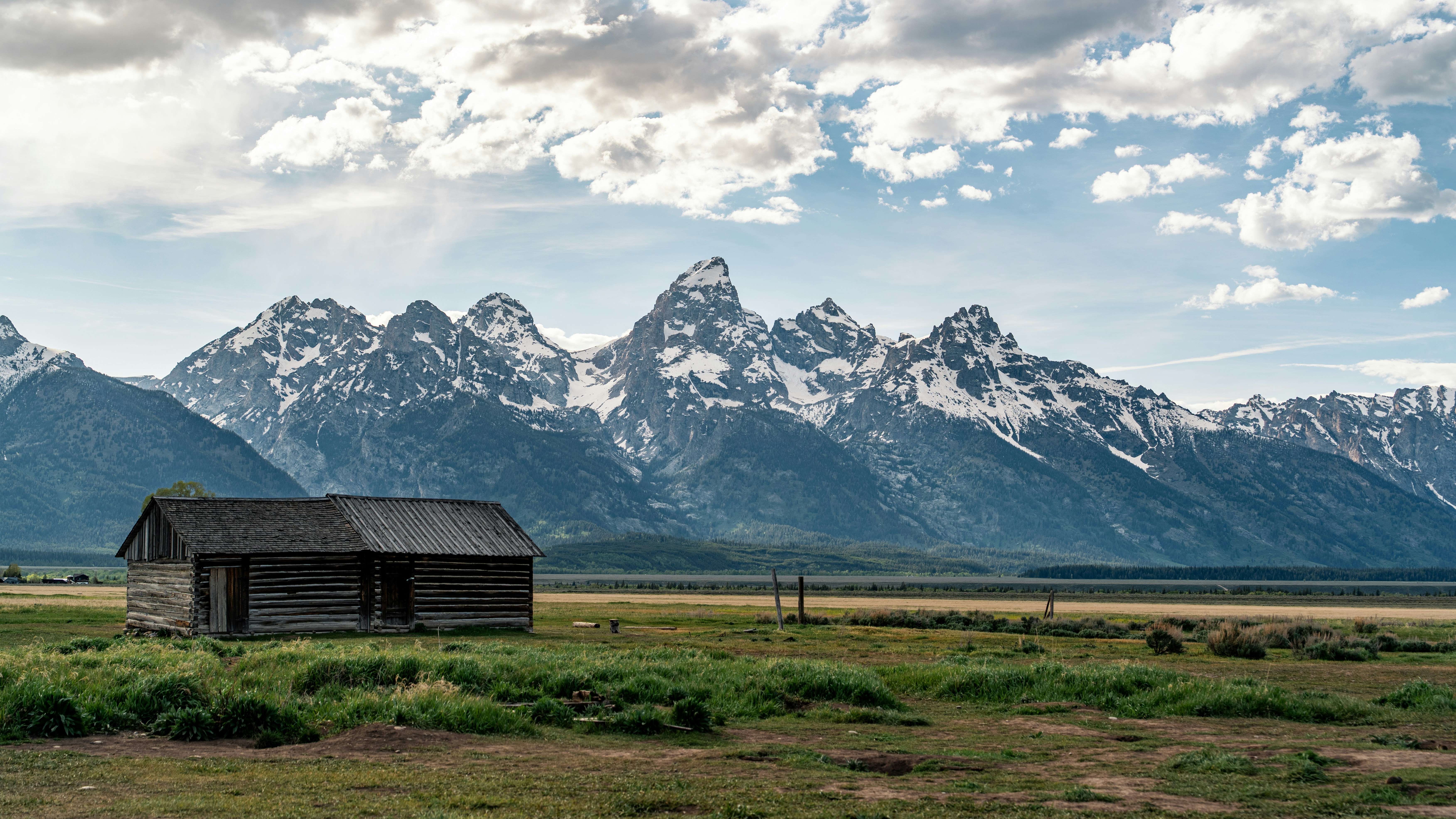 A rustic log cabin in a grassy field before the snow-capped Teton Range