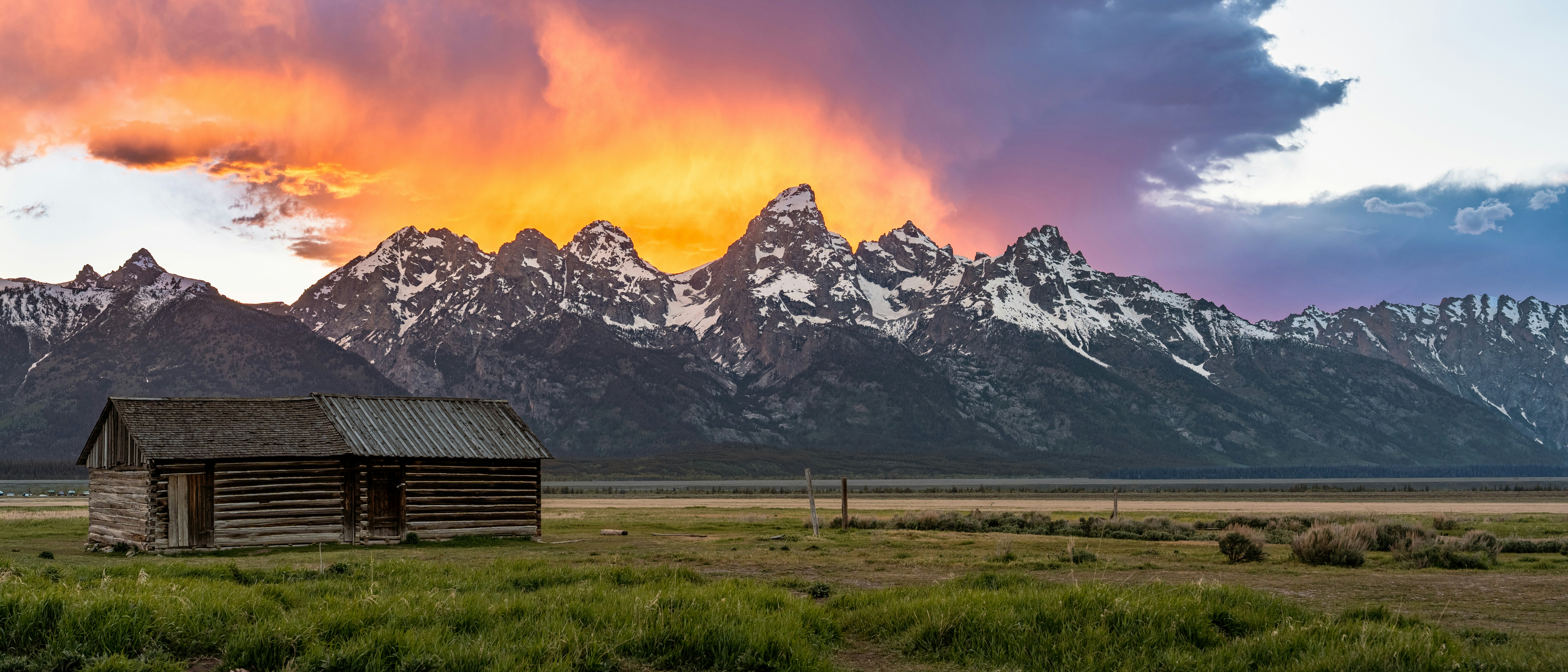 A rustic log cabin in a grassy field before snow-capped mountains under a glowing sky