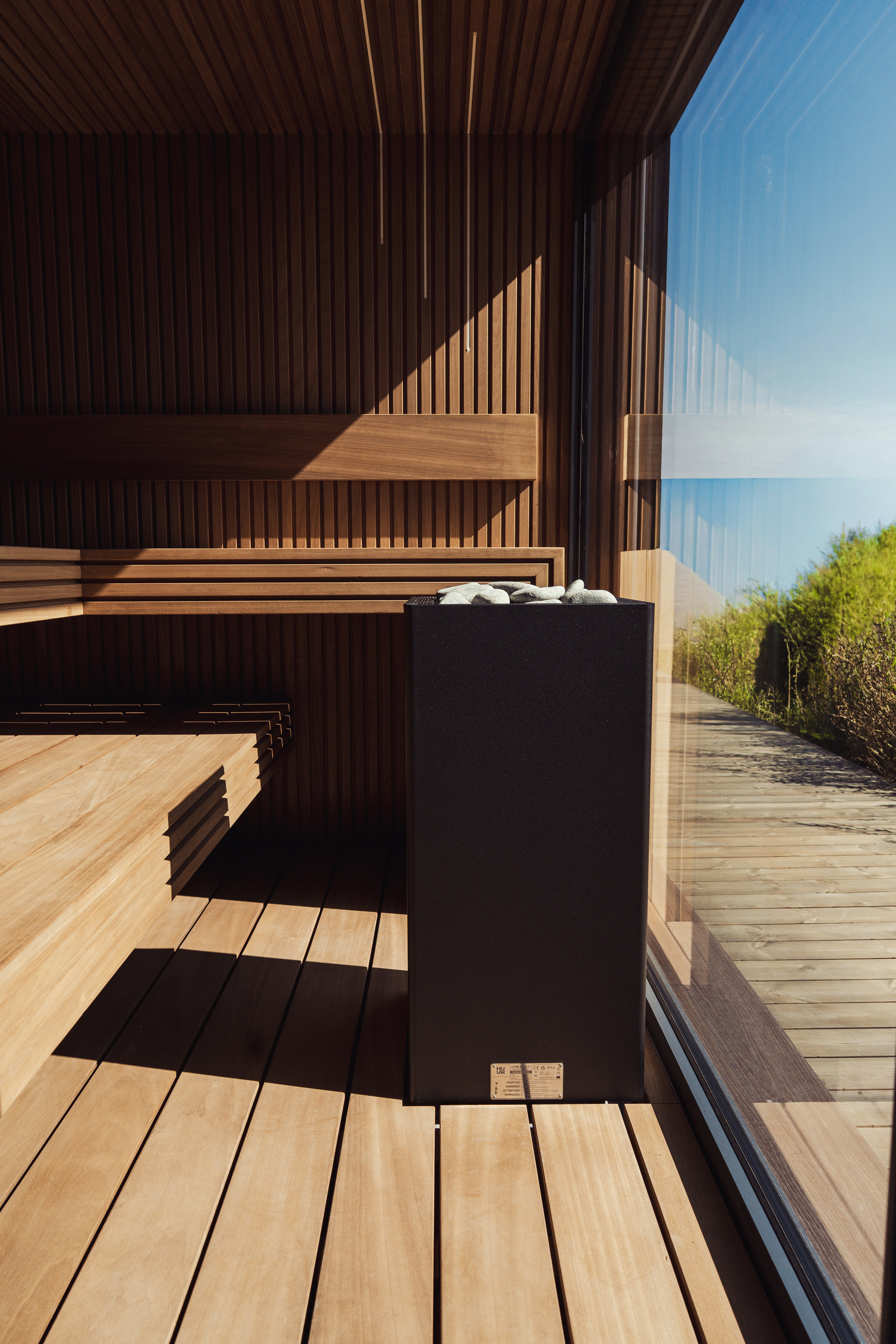 Um aquecedor de sauna preto com pedras dentro de uma sauna de madeira com paredes de vidro