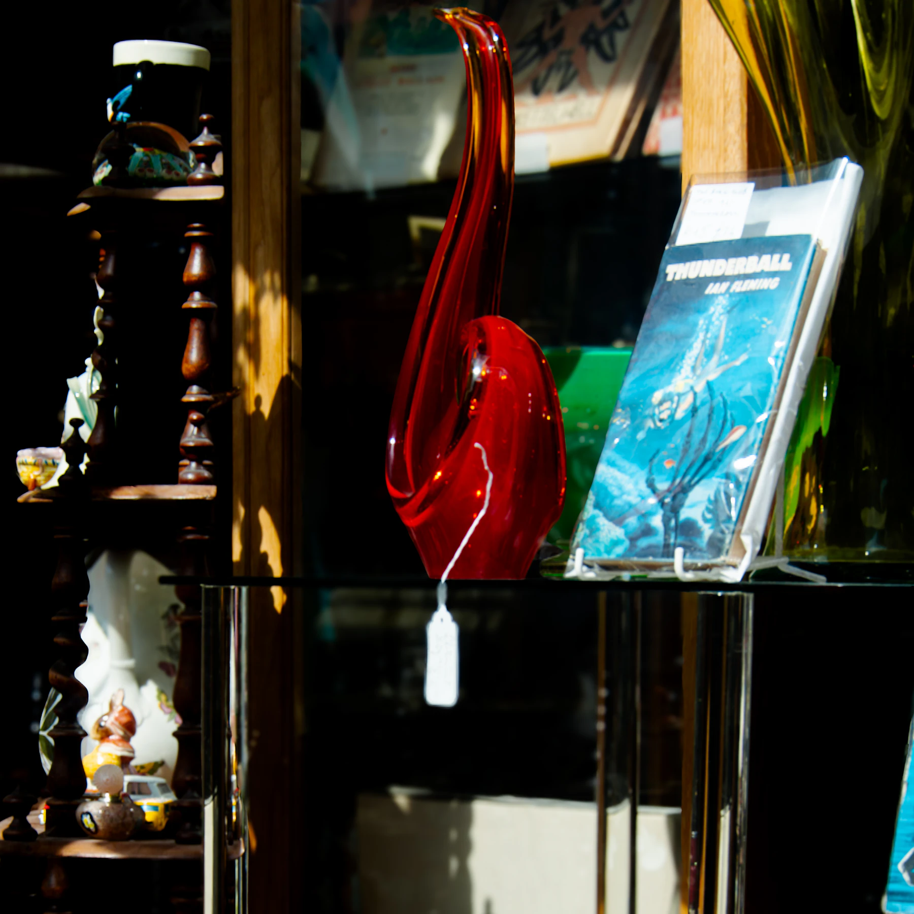 A red art-glass sculpture with a price tag beside a paperback book on a glass shelf in a shop