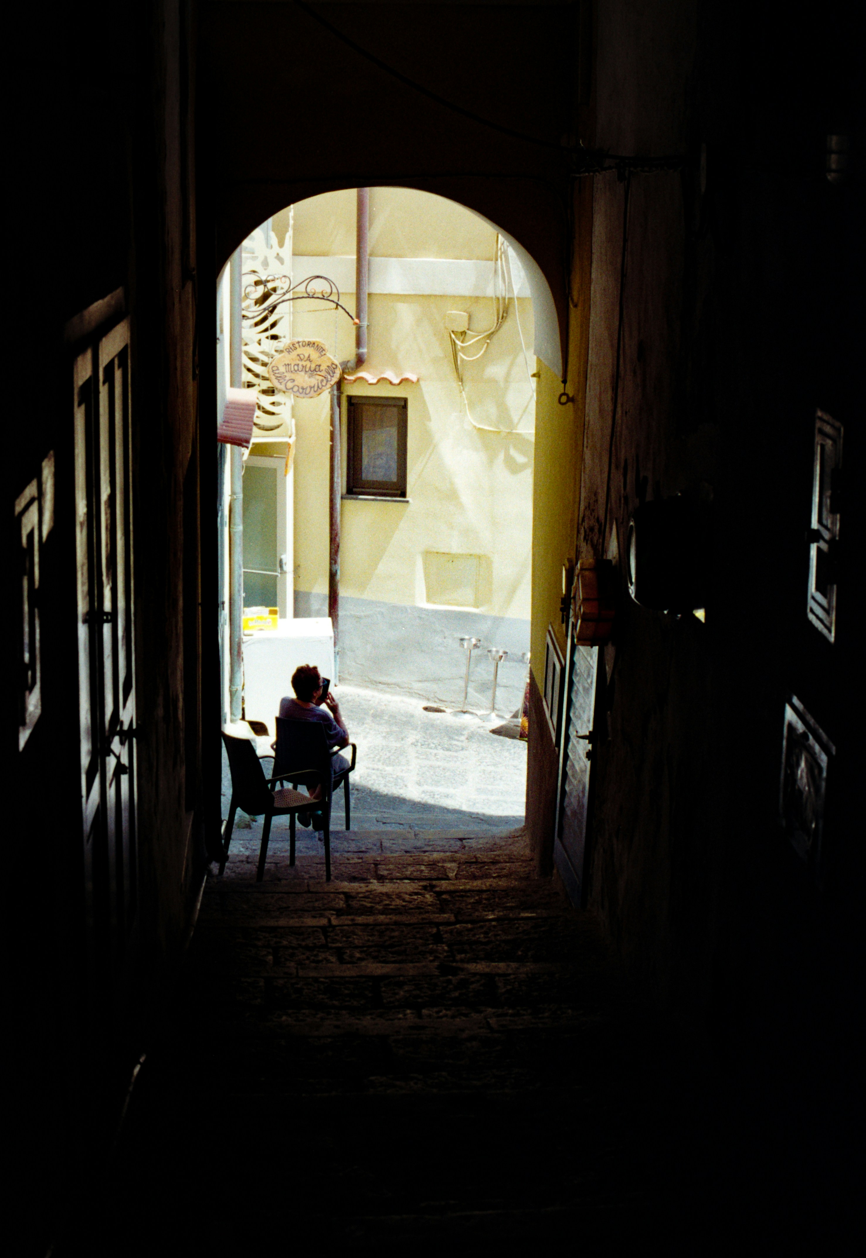 Una persona sentada en una silla en un callejón iluminado por el sol vista desde un pasadizo oscuro