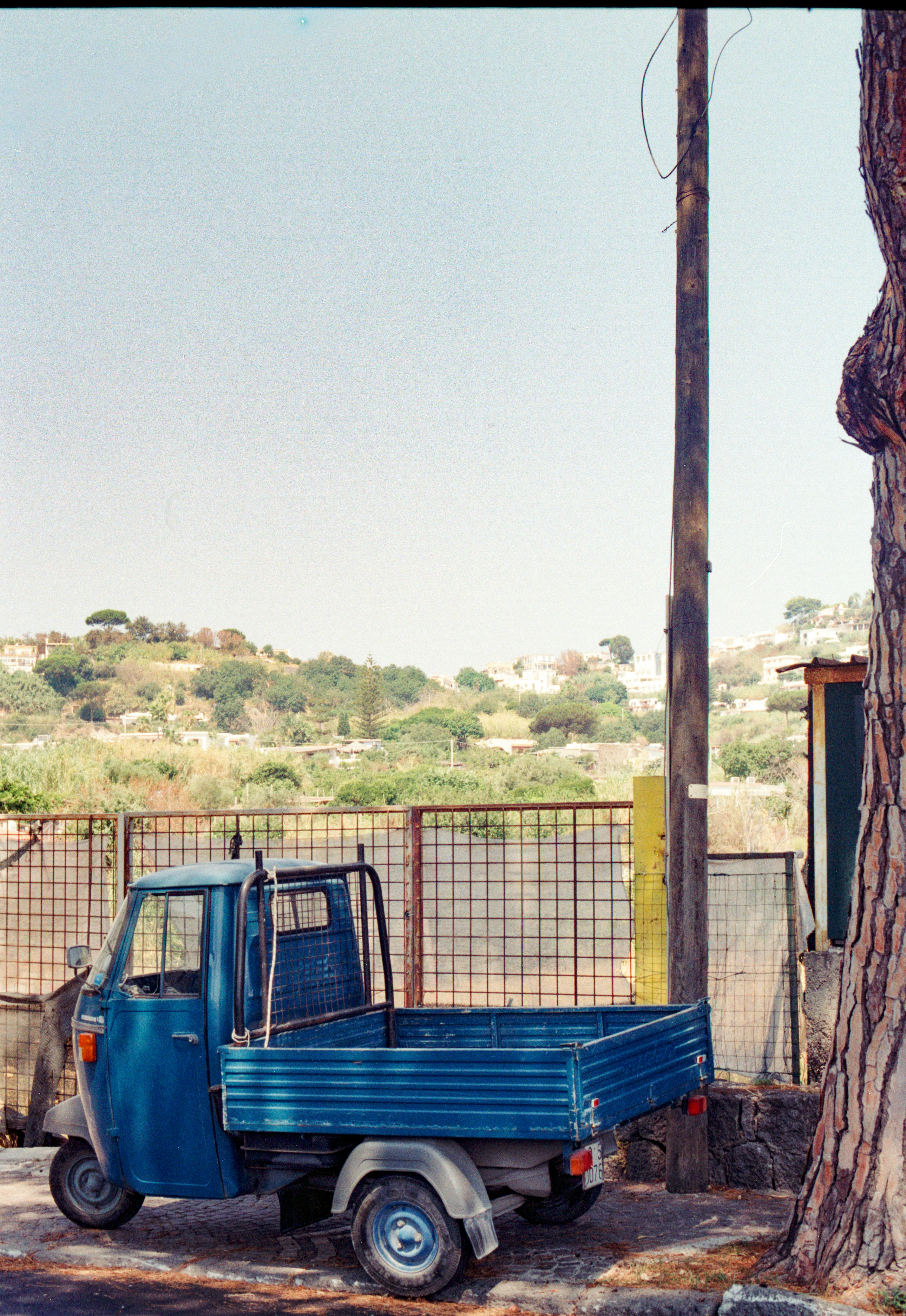 Un camión Piaggio Ape azul de tres ruedas aparcado junto a un poste de madera,