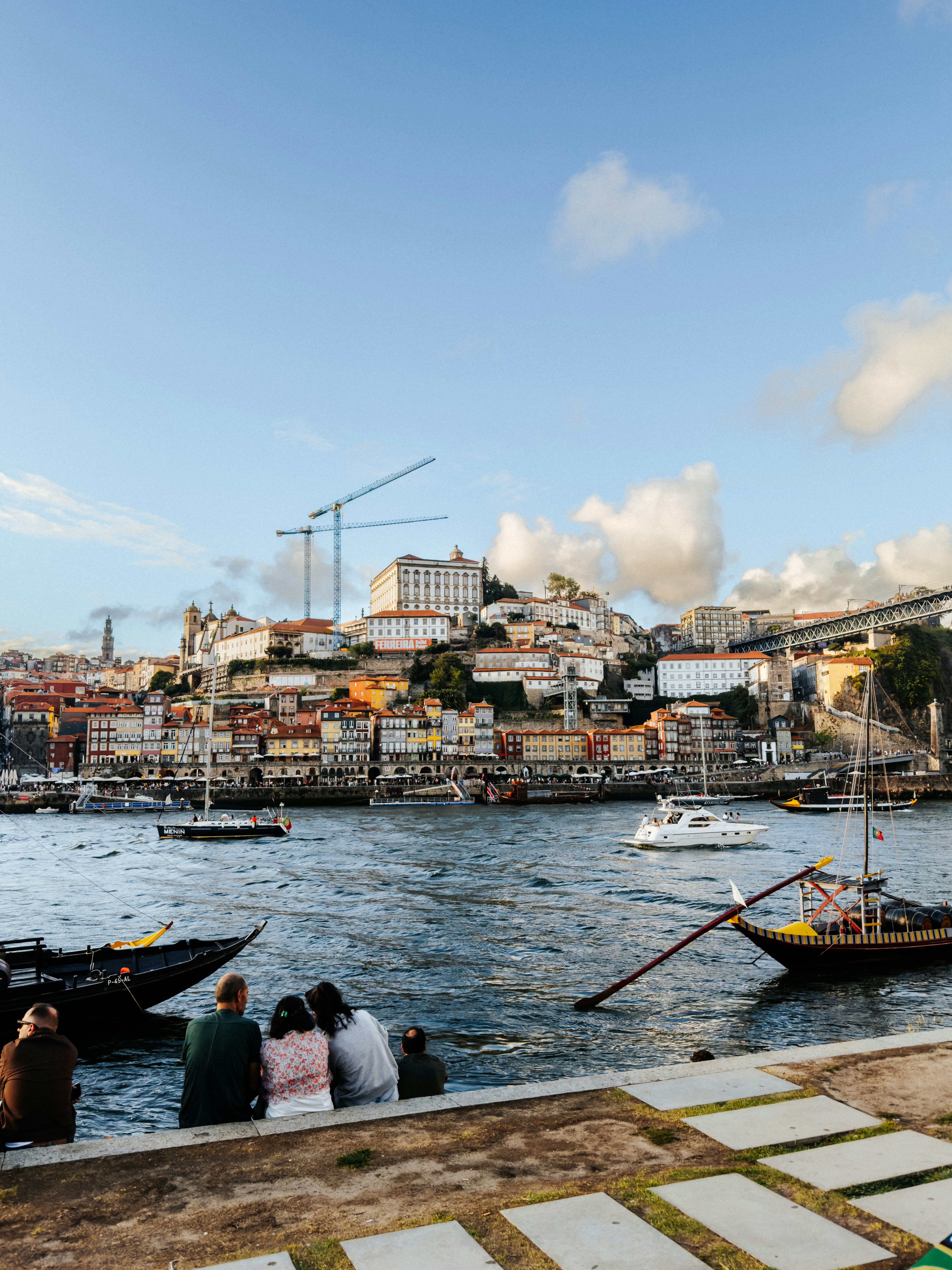People sitting on a riverbank in Porto with traditional boats and city buildings