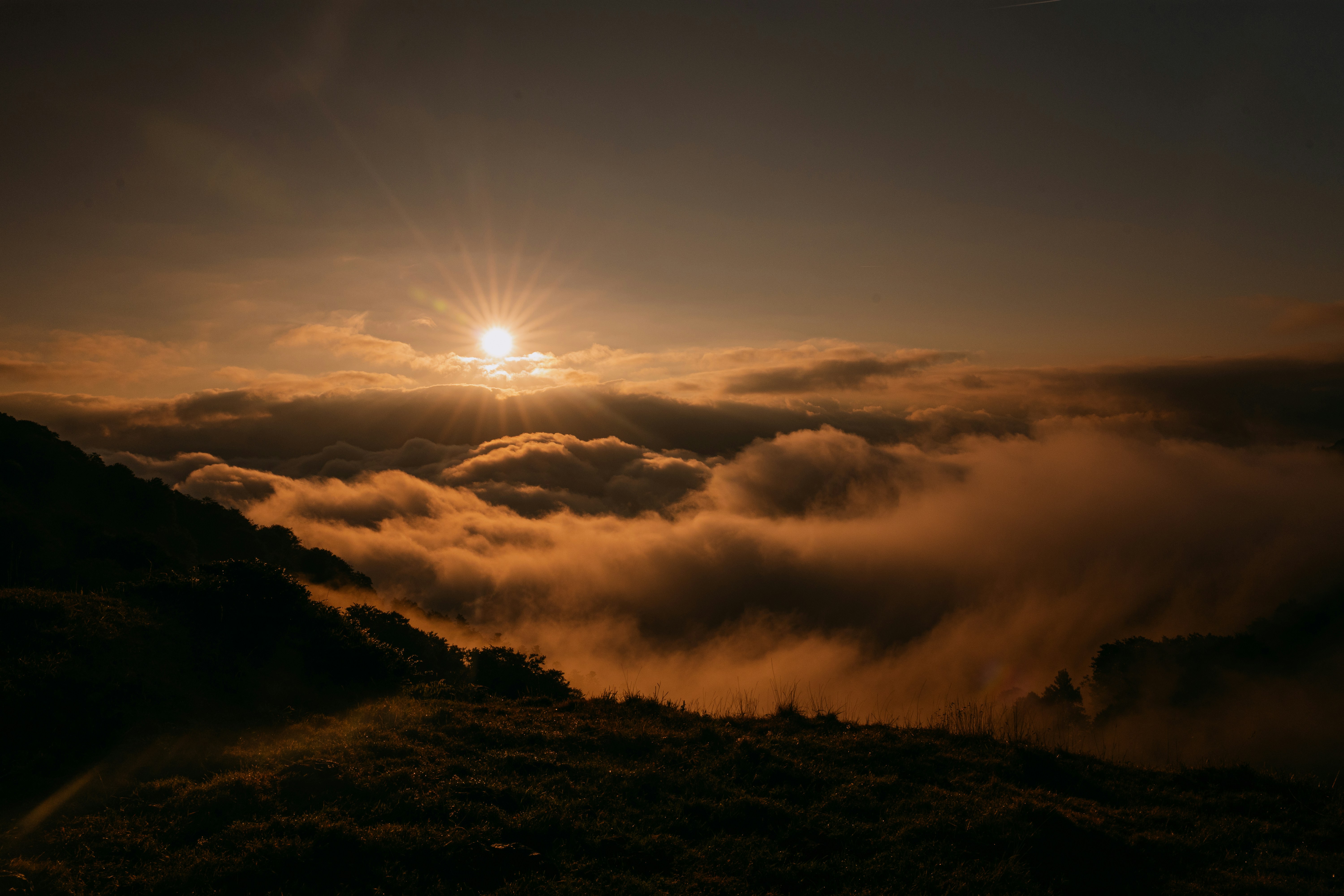 Low sun shining over a thick layer of clouds above a dark hillside