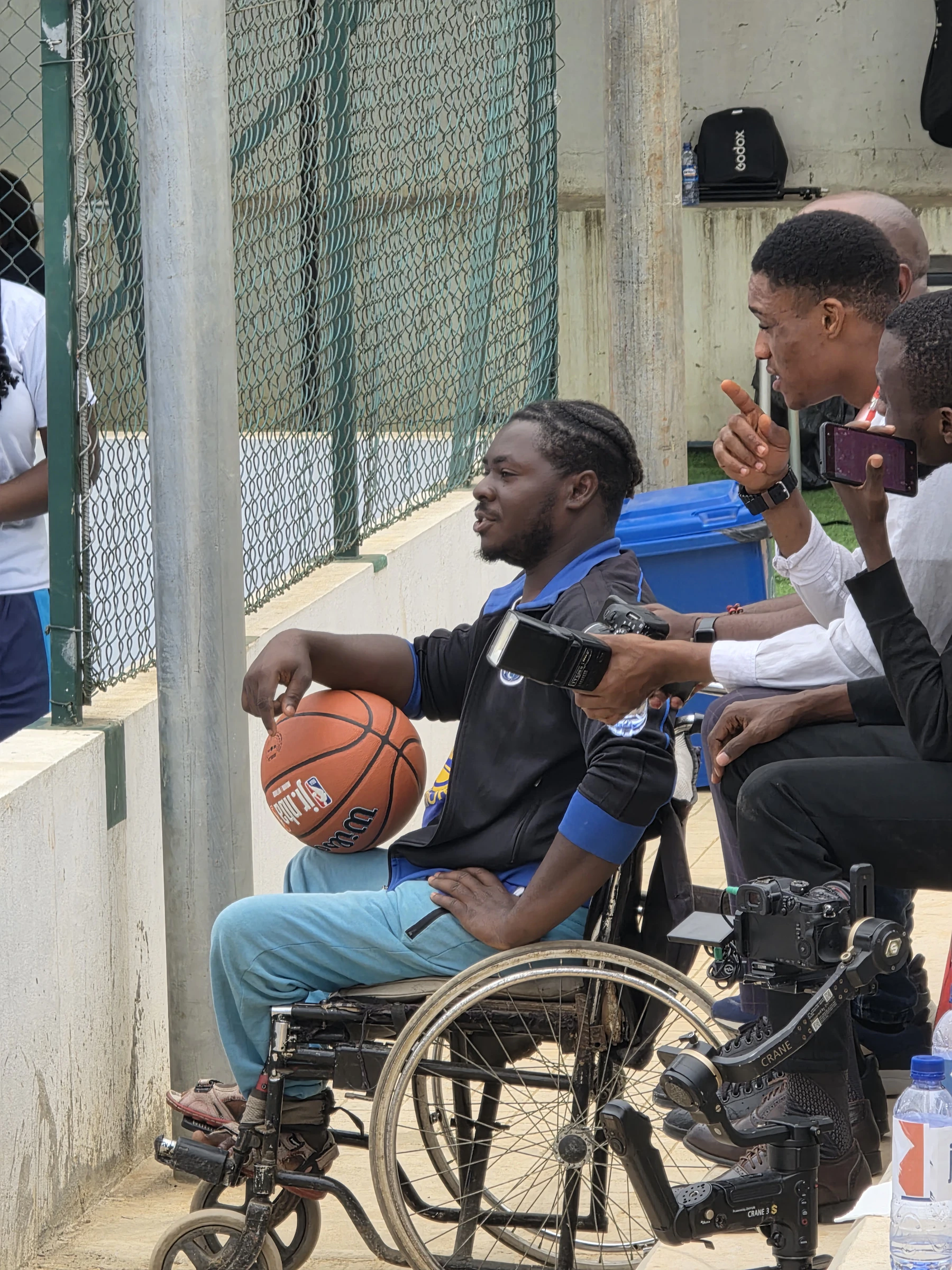 A man in an everyday wheelchair holding a basketball at the side of a fenced outdoor court