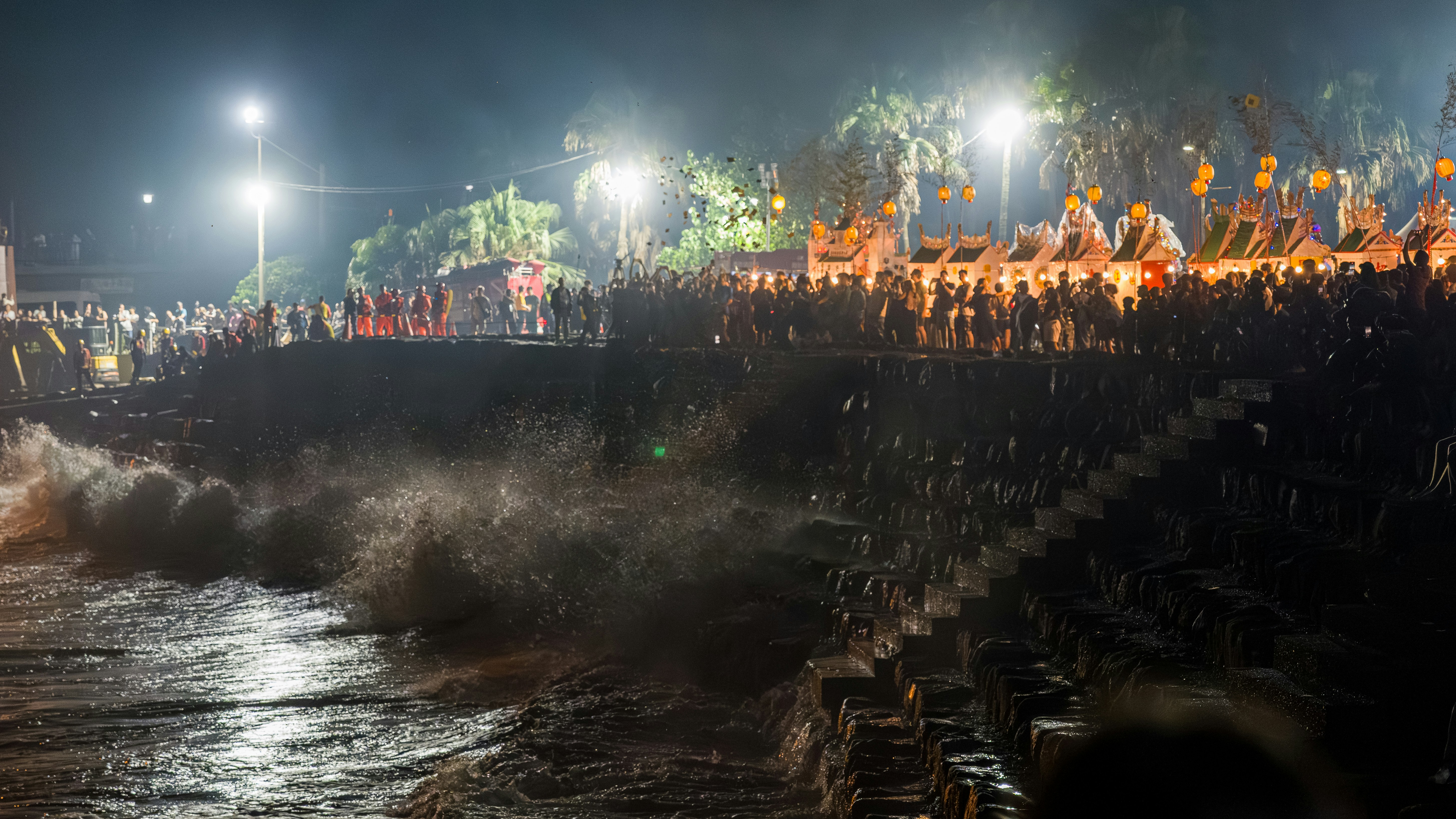 A crowd of people watching waves crash against a stone embankment at night