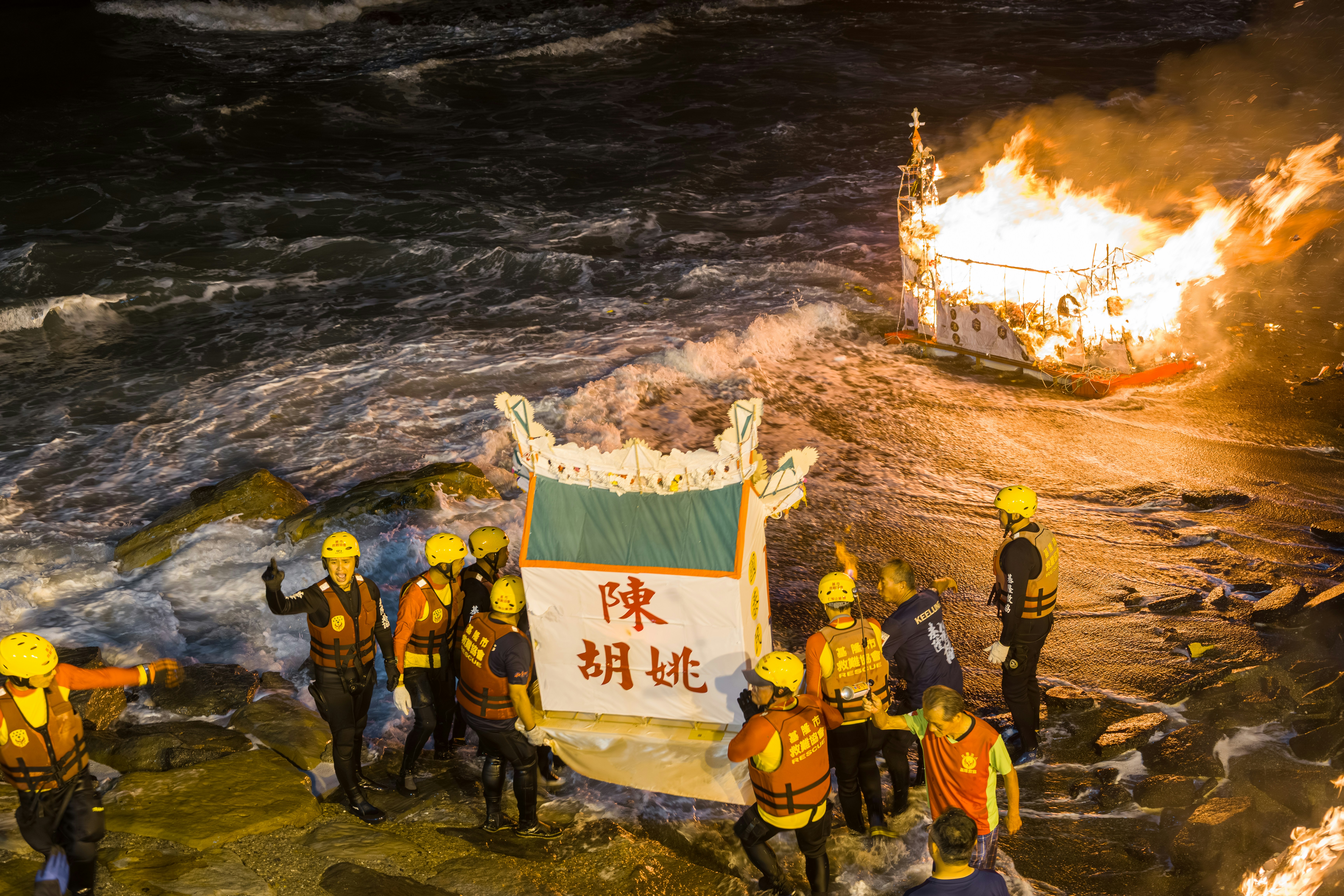 Rescue workers in life vests carrying a ceremonial paper structure on a rocky beach