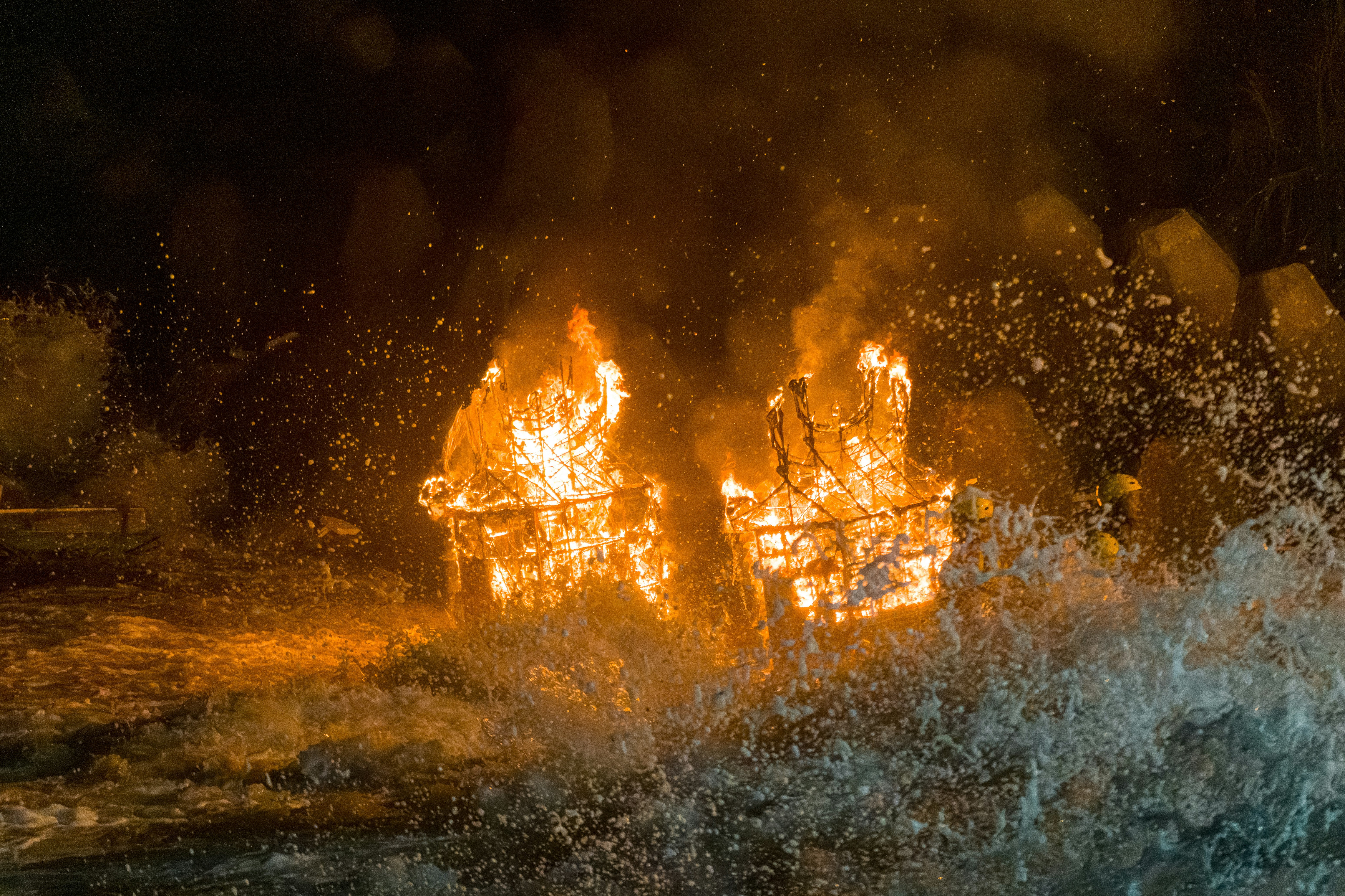 Two burning wooden structures engulfed in flames amidst splashing sea water at night
