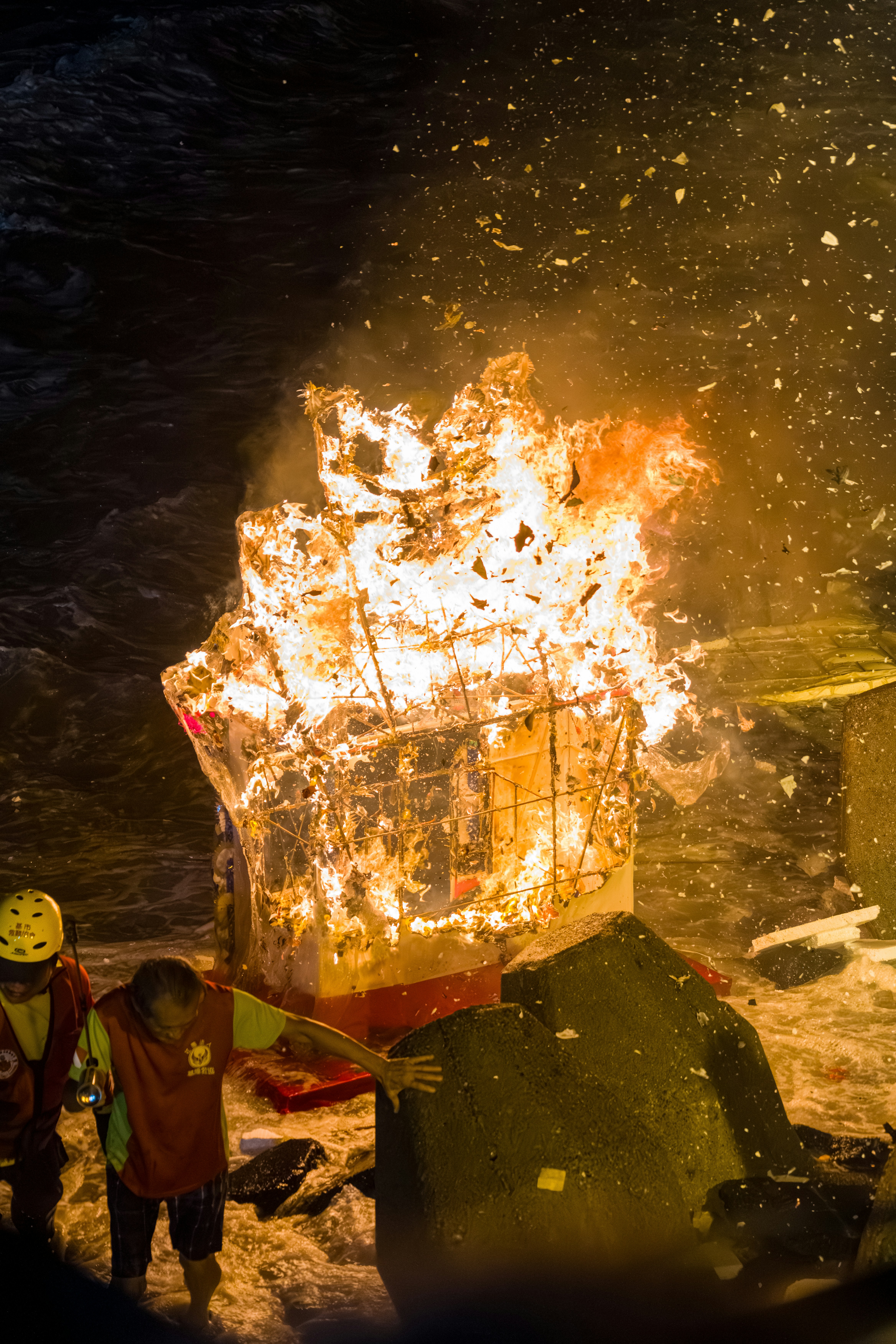 People in safety gear near a burning structure on a dark rocky shore