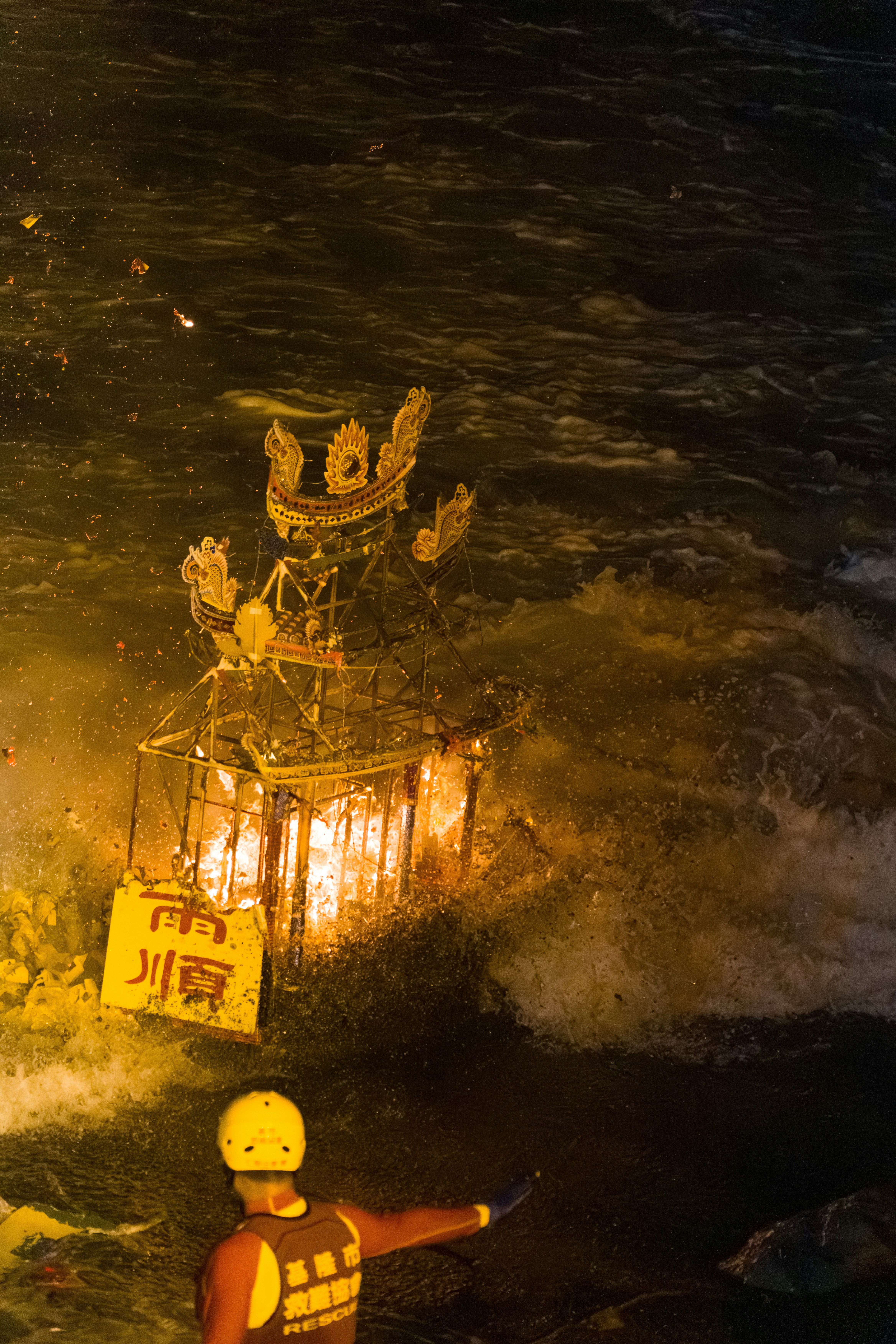A burning ornate structure in dark water with a rescuer in the foreground