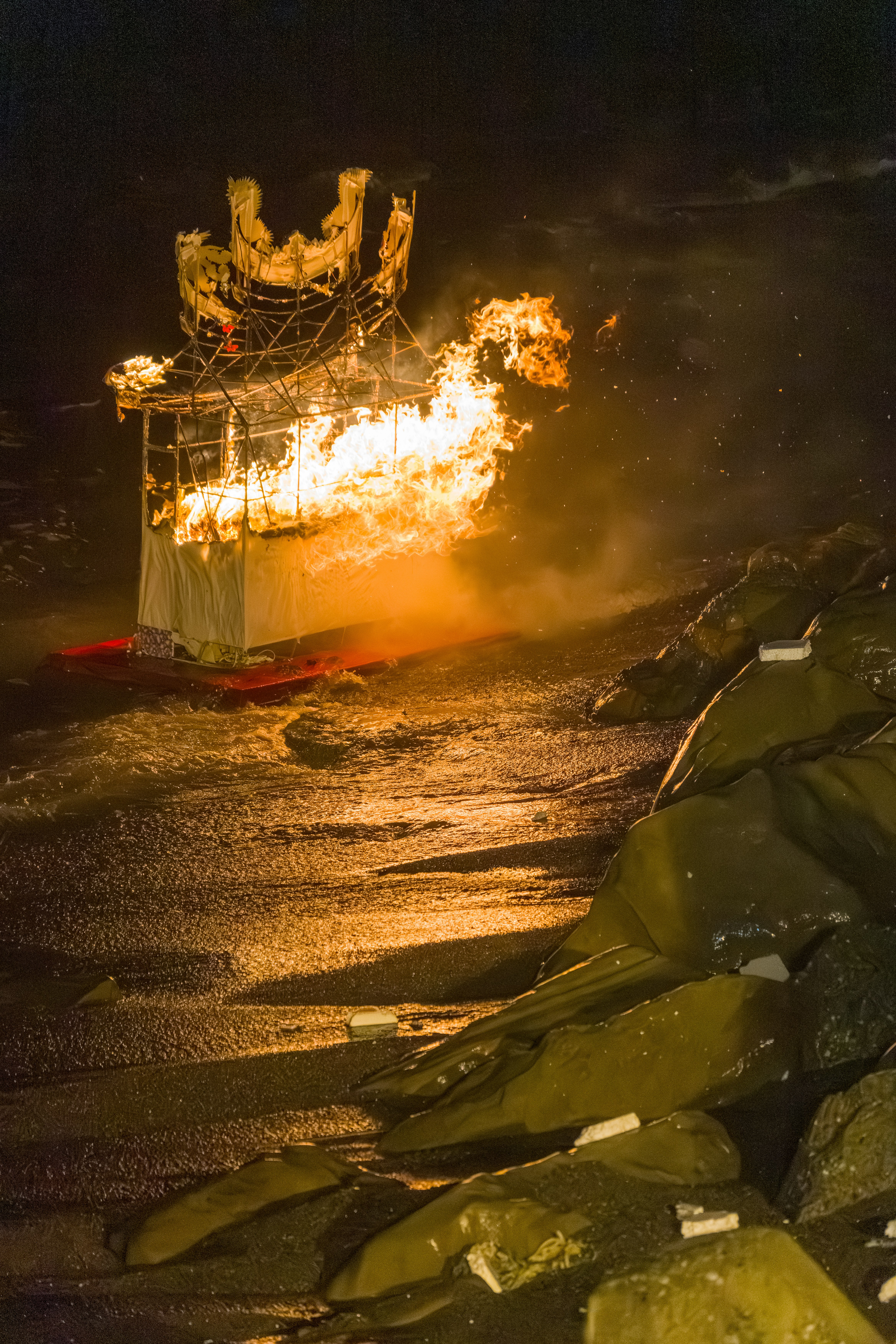 A burning structure on a red platform floating in dark water near rocks