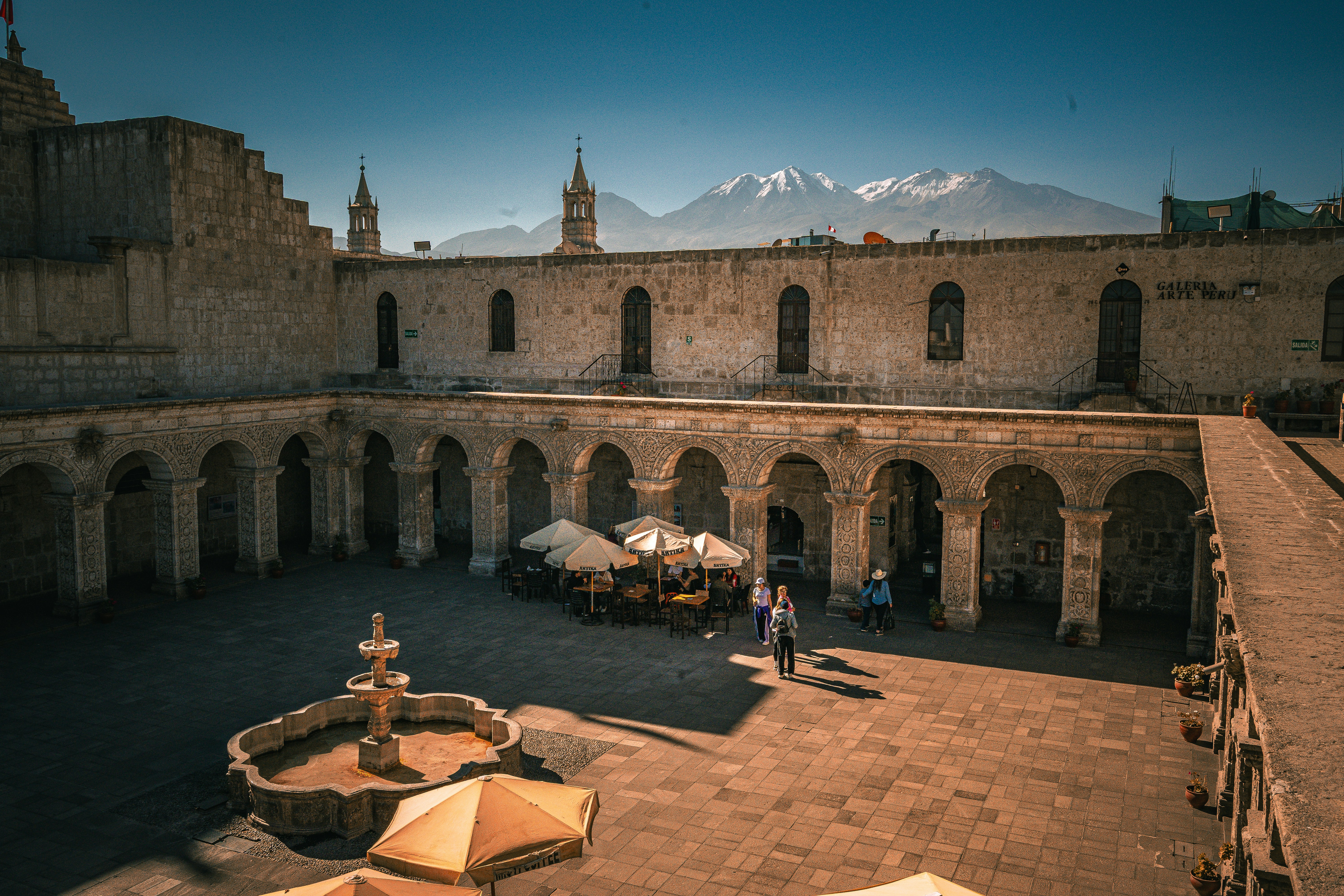 A stone courtyard with a fountain and cafe tables beneath snow-capped mountains in Arequipa