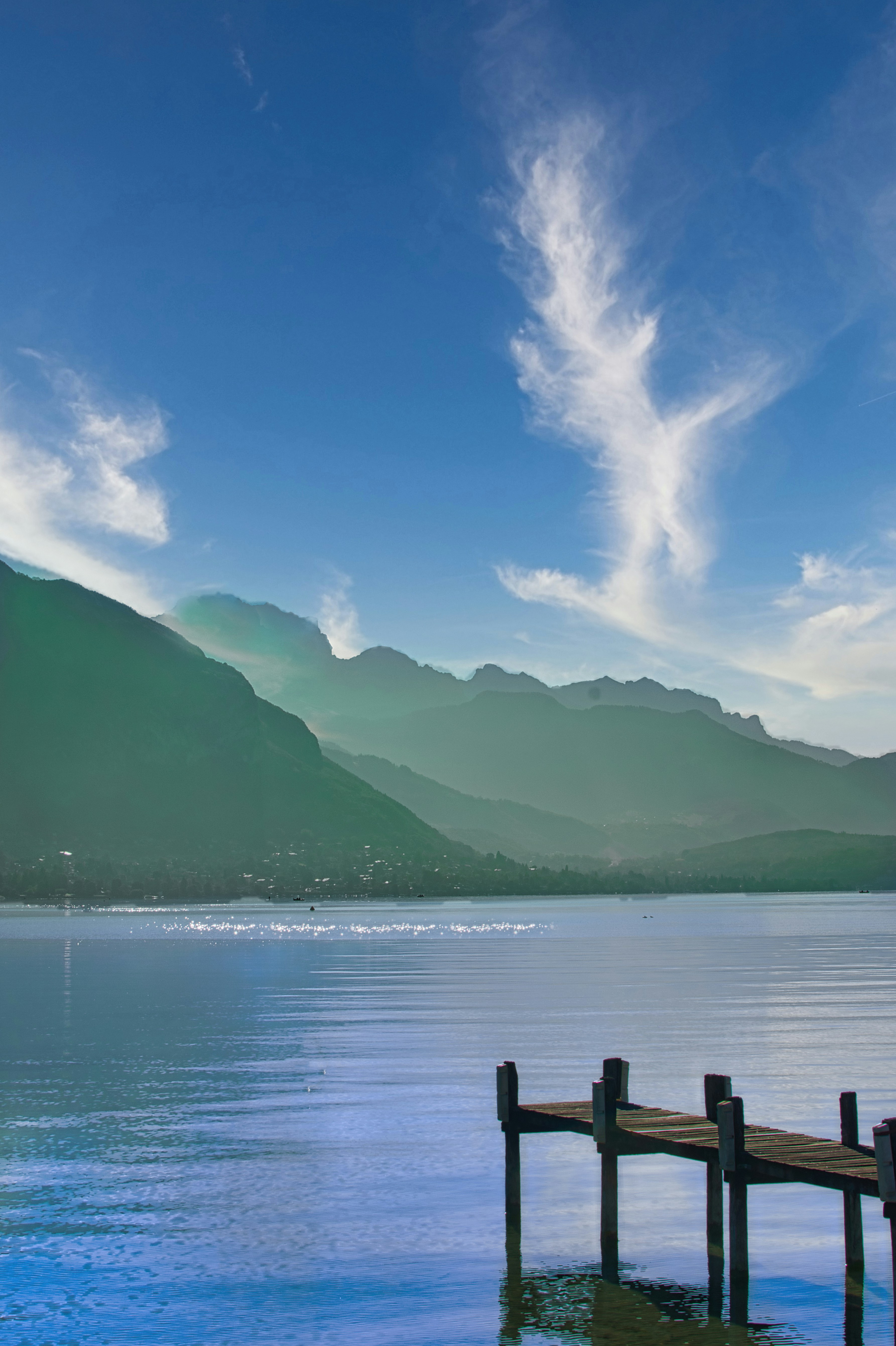 A wooden pier extending into a calm lake beneath distant mountains and wispy clouds